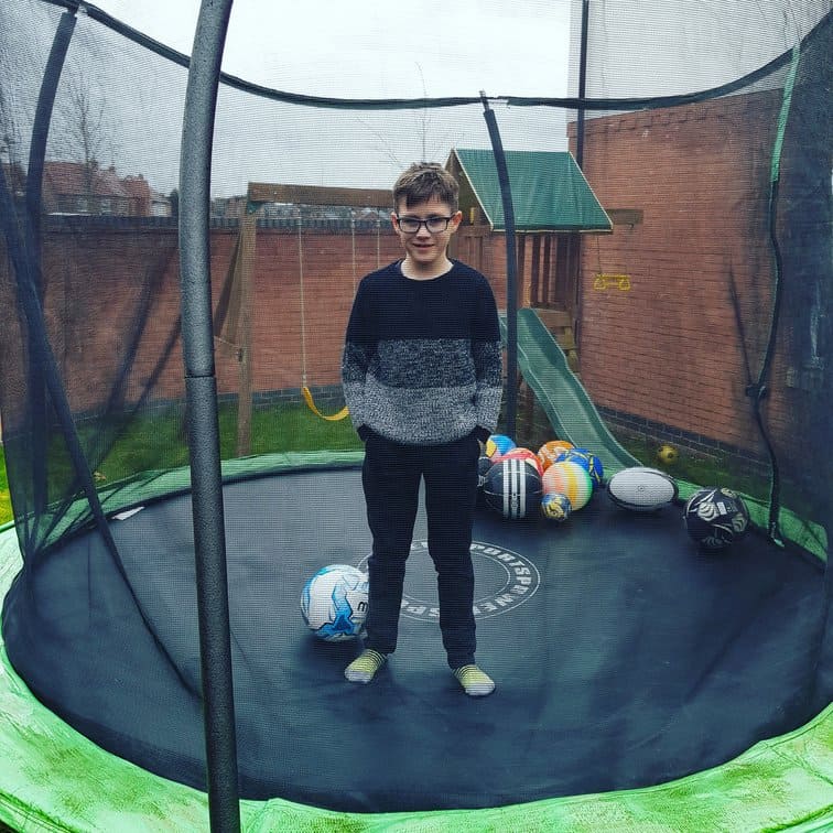 young boy standing on a trampoline with basketballs, soccer balls, and rugby balls to play with