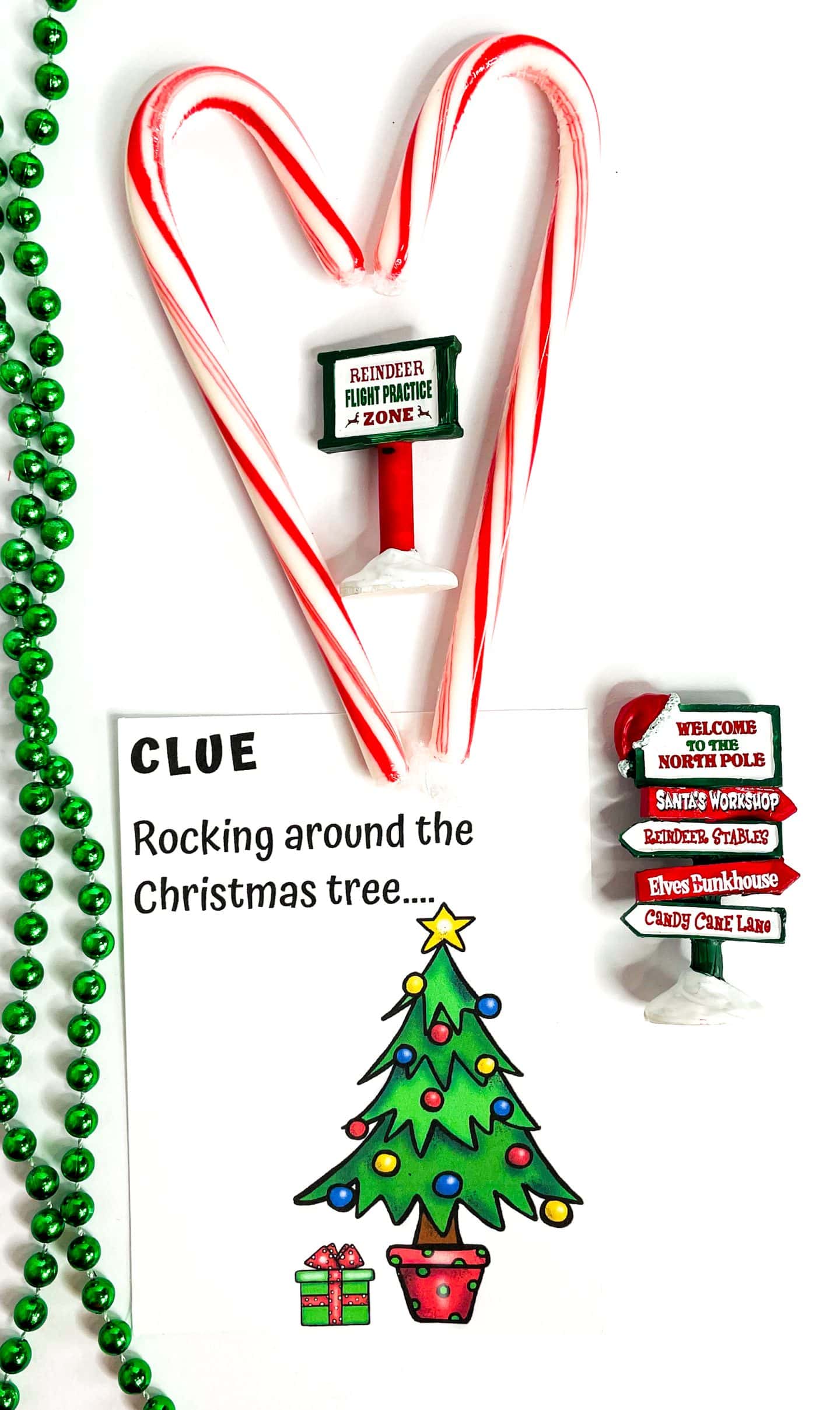A Christmas scavenger hunt clue card reading “Rocking around the Christmas tree…” lies next to festive props including two candy canes arranged in a heart shape, a “Reindeer Flight Practice Zone” sign, a green beaded necklace, and a North Pole-themed signpost pointing to Santa’s Workshop and Candy Cane Lane. The clue card features a decorated Christmas tree and a wrapped gift, making it a playful and colorful addition to holiday scavenger hunt fun.

