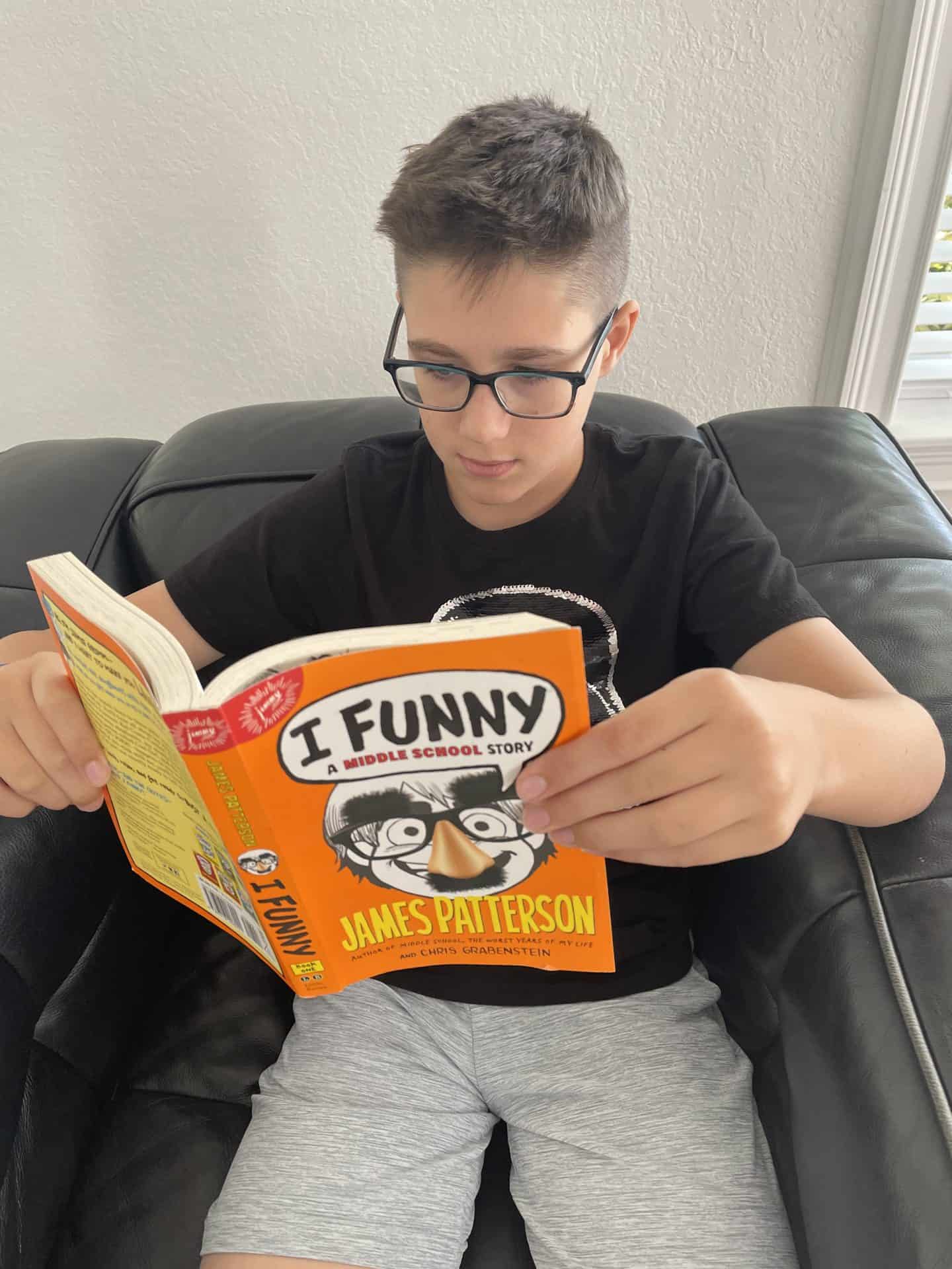 A boy wearing glasses and a black T-shirt sits on a black leather couch reading the book I Funny: A Middle School Story by James Patterson. The book cover is bright orange and yellow, featuring a cartoon face with a fake nose, glasses, and mustache.