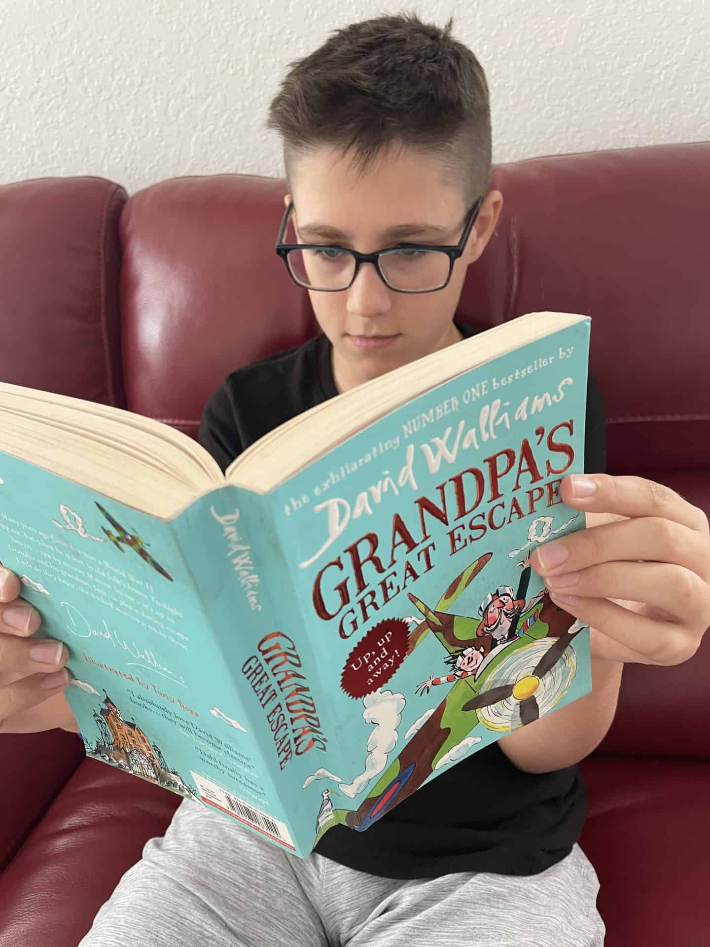 A boy with glasses sits on a red couch reading the book Grandpa’s Great Escape by David Walliams. The book cover is teal and illustrated with a cartoonish scene of a grandpa and child flying a vintage green airplane over a whimsical landscape.