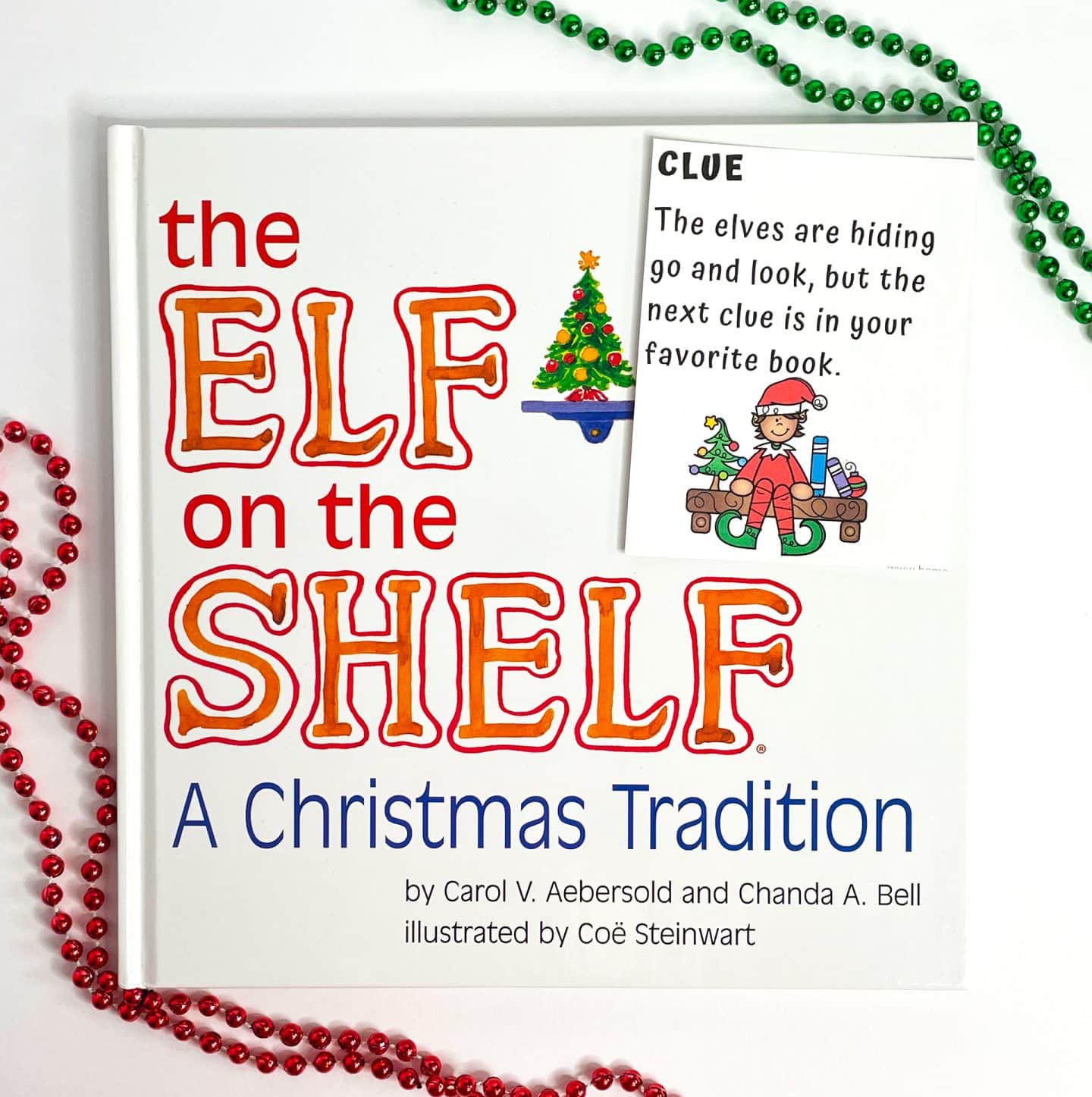 A copy of The Elf on the Shelf: A Christmas Tradition book is displayed with festive red and green bead garlands. On top of the book sits one of the elf on the shelf scavenger hunt clues, reading “The elves are hiding go and look, but the next clue is in your favorite book,” with an illustration of a cheerful elf surrounded by toys and candy canes. This setup highlights a fun holiday reading and scavenger hunt activity for kids.


