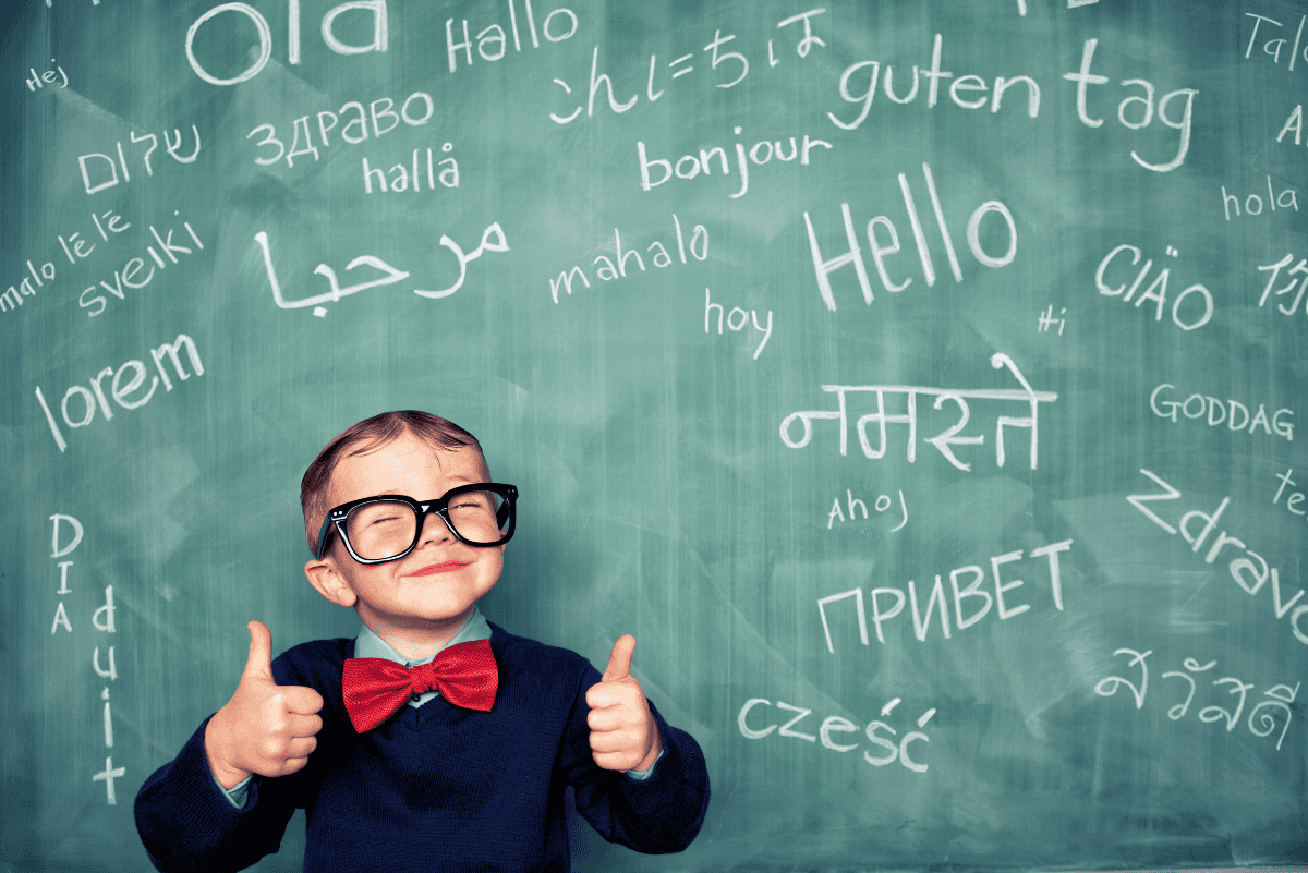 Smiling young boy in glasses and a red bow tie stands in front of a chalkboard covered with the word “Hello” written in multiple languages. With both thumbs up, he represents enthusiasm for learning languages—an ideal visual for promoting the best foreign language curriculum for homeschoolers.