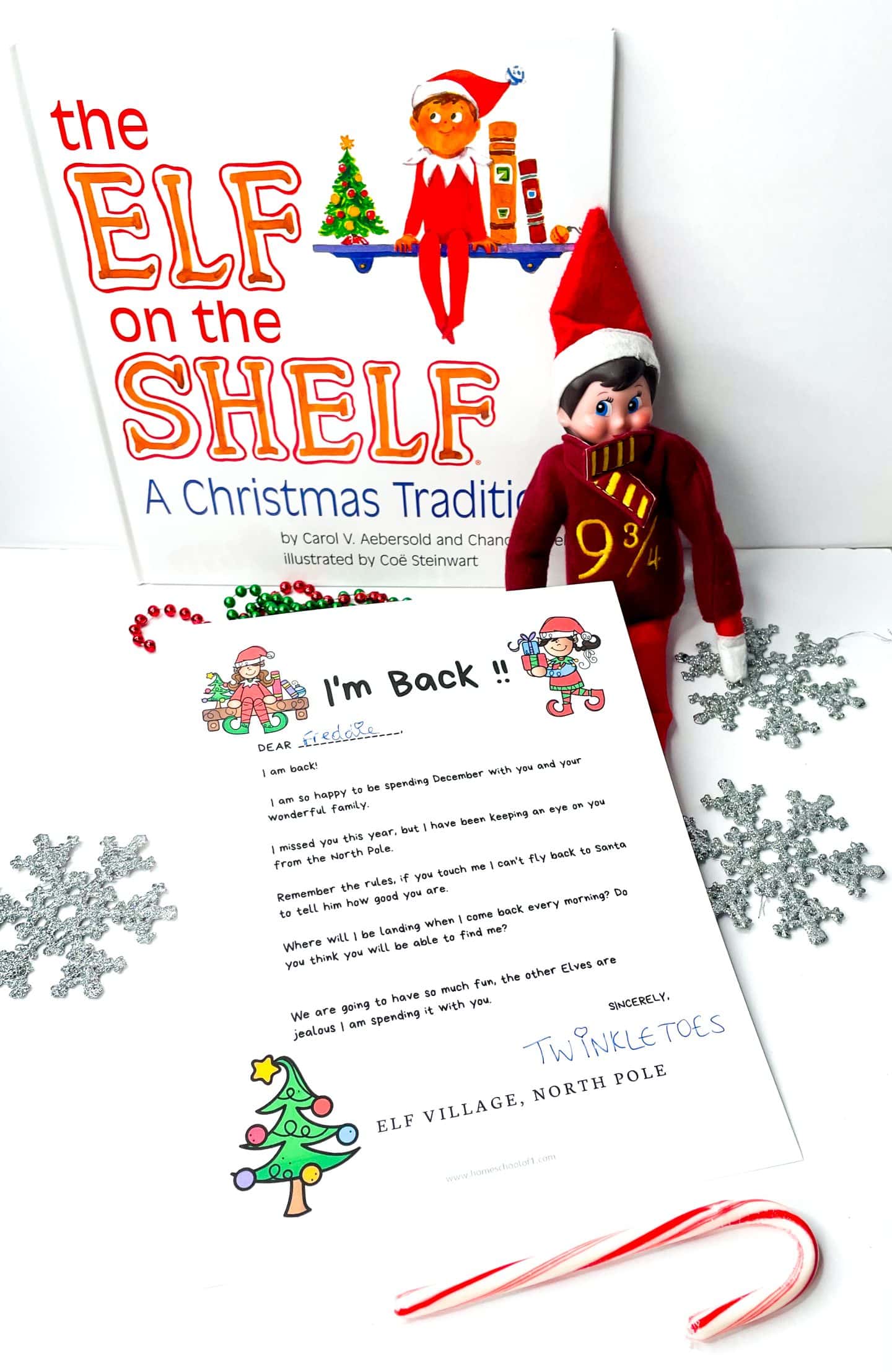 An elf on the shelf arrival letter titled “I’m Back!!” is placed in front of The Elf on the Shelf: A Christmas Tradition book, alongside a festive elf doll in a Harry Potter-inspired outfit. The letter, addressed to Freddie and signed by “Twinkle Toes,” welcomes the child to a fun December with reminders about the elf rules. Surrounded by red and green bead garlands, glittery snowflakes, and a candy cane, this cheerful elf on the shelf arrival letter adds excitement to the Christmas countdown.

