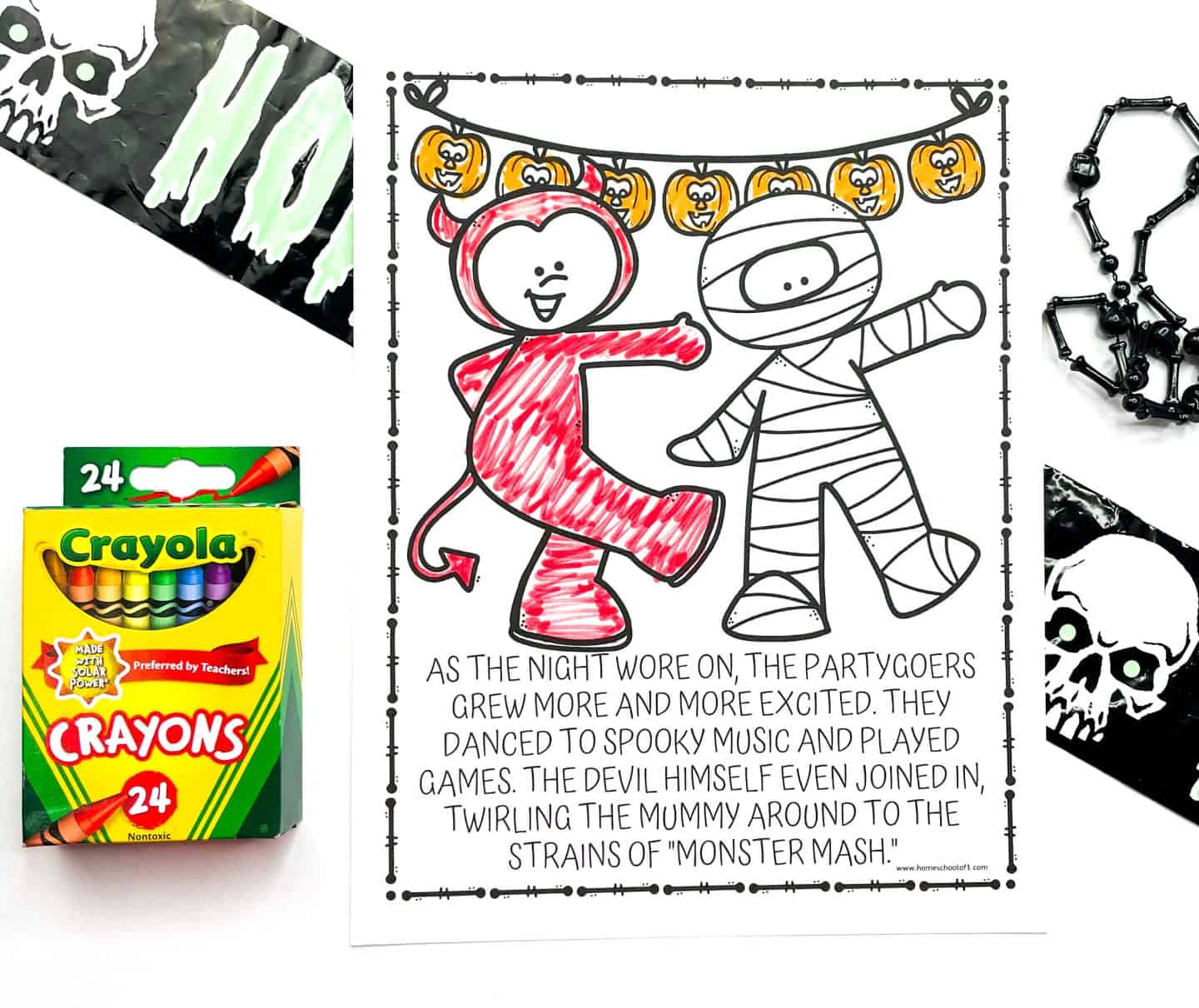 A Halloween coloring page shows a cartoon devil and mummy dancing under a string of jack-o’-lantern lights. The devil character is colored in red, while the rest of the page is partially uncolored. Below the image is a short story mentioning a spooky party and the song “Monster Mash.” The scene is decorated with Halloween props including a box of Crayola crayons, a black skull-themed banner, and a plastic chain, creating a festive and playful setup.