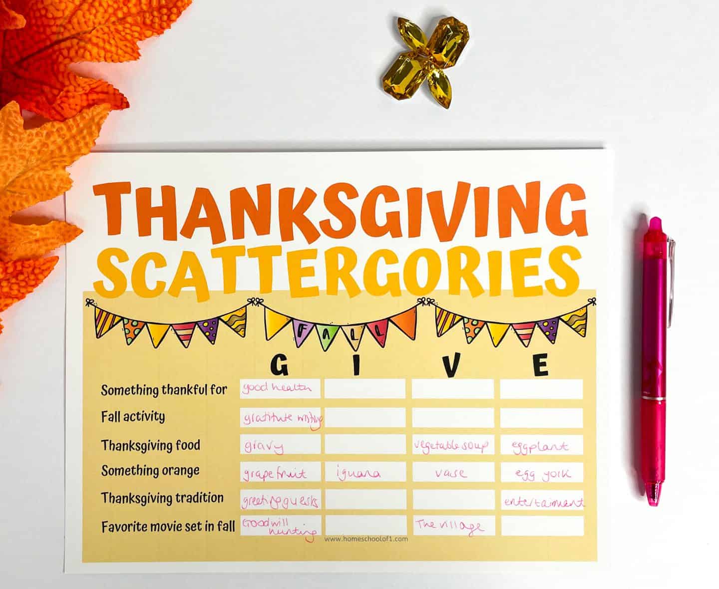 A completed Thanksgiving Scattergories game card titled “GIVE,” featuring categories like "Thanksgiving food" and "Fall activity" with handwritten answers in pink ink. Surrounded by festive fall decor, a pink pen, and orange leaves, this printable game is perfect for holiday fun.