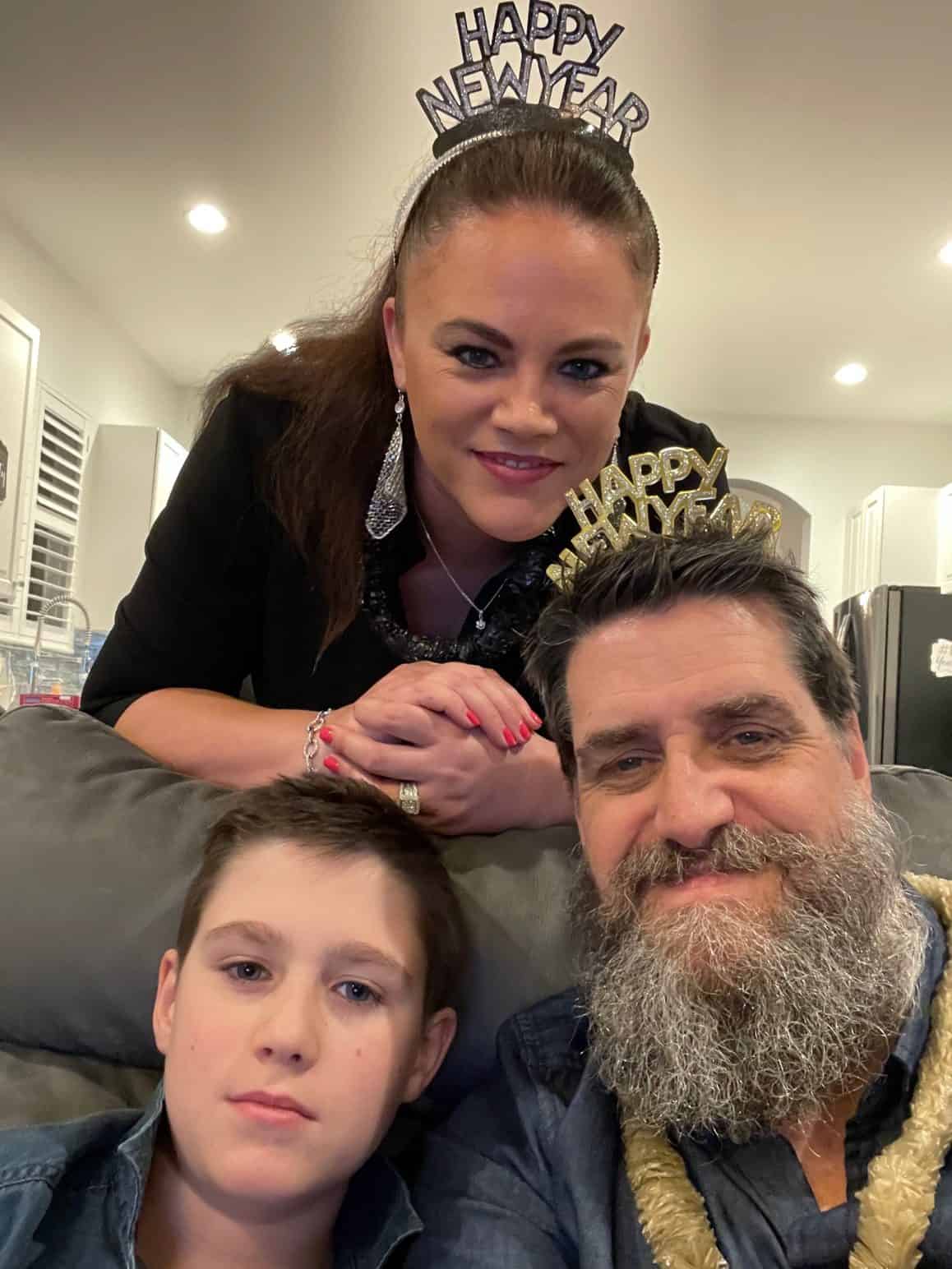 Three people celebrate New Year’s Eve indoors, smiling and dressed up with “Happy New Year” headbands. The woman stands behind the couch wearing sparkly earrings and makeup, while the bearded man and boy sit in front. They’re surrounded by warm lighting and festive vibes in a cozy home setting.

