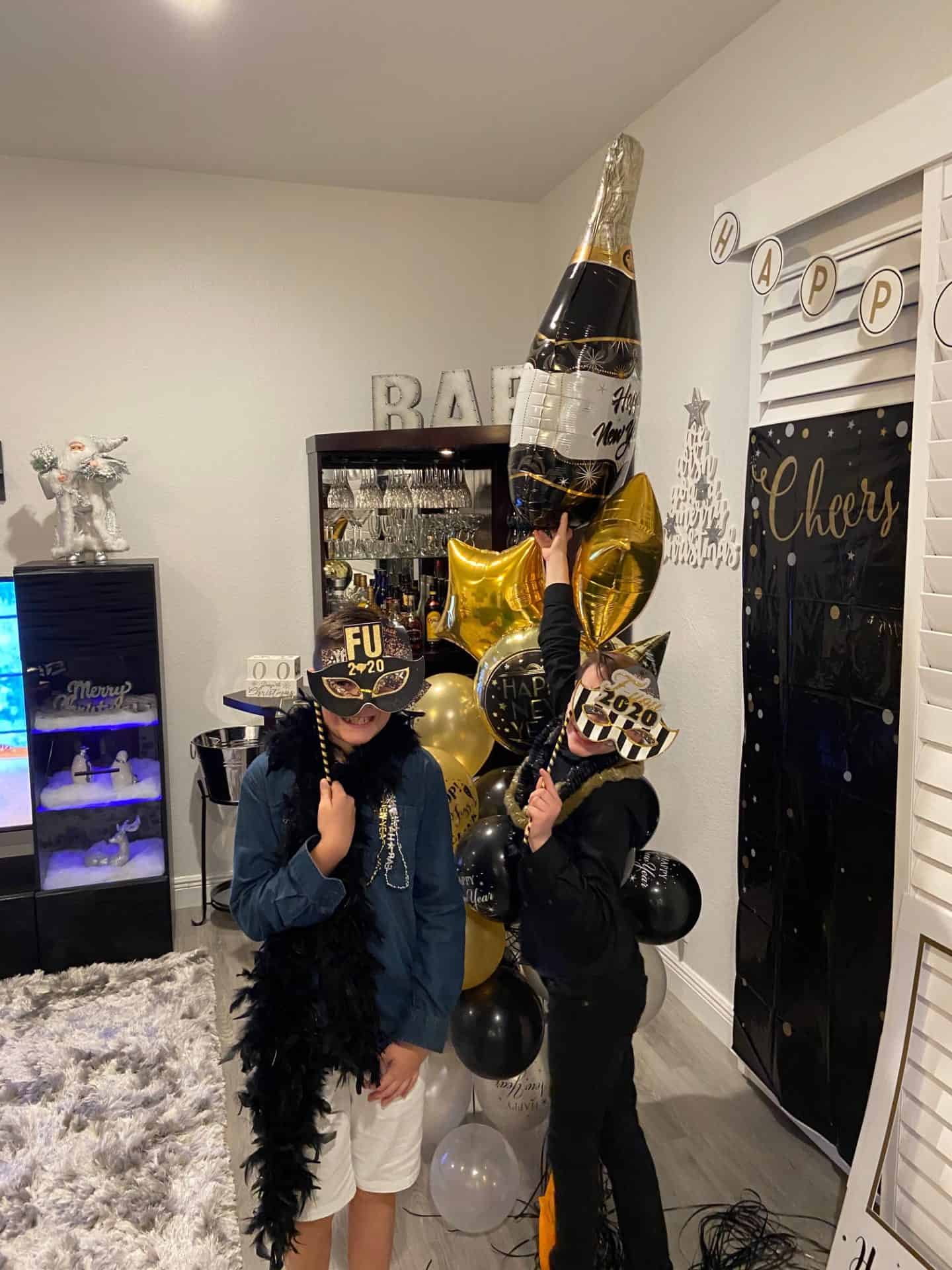 Two kids celebrate New Year's Eve at home, wearing festive hats that say "FU 2020" and posing with a large champagne bottle balloon surrounded by black, gold, and silver balloons. The room is decorated with New Year’s banners, a “Cheers” backdrop, and a home bar setup, creating a fun party atmosphere.