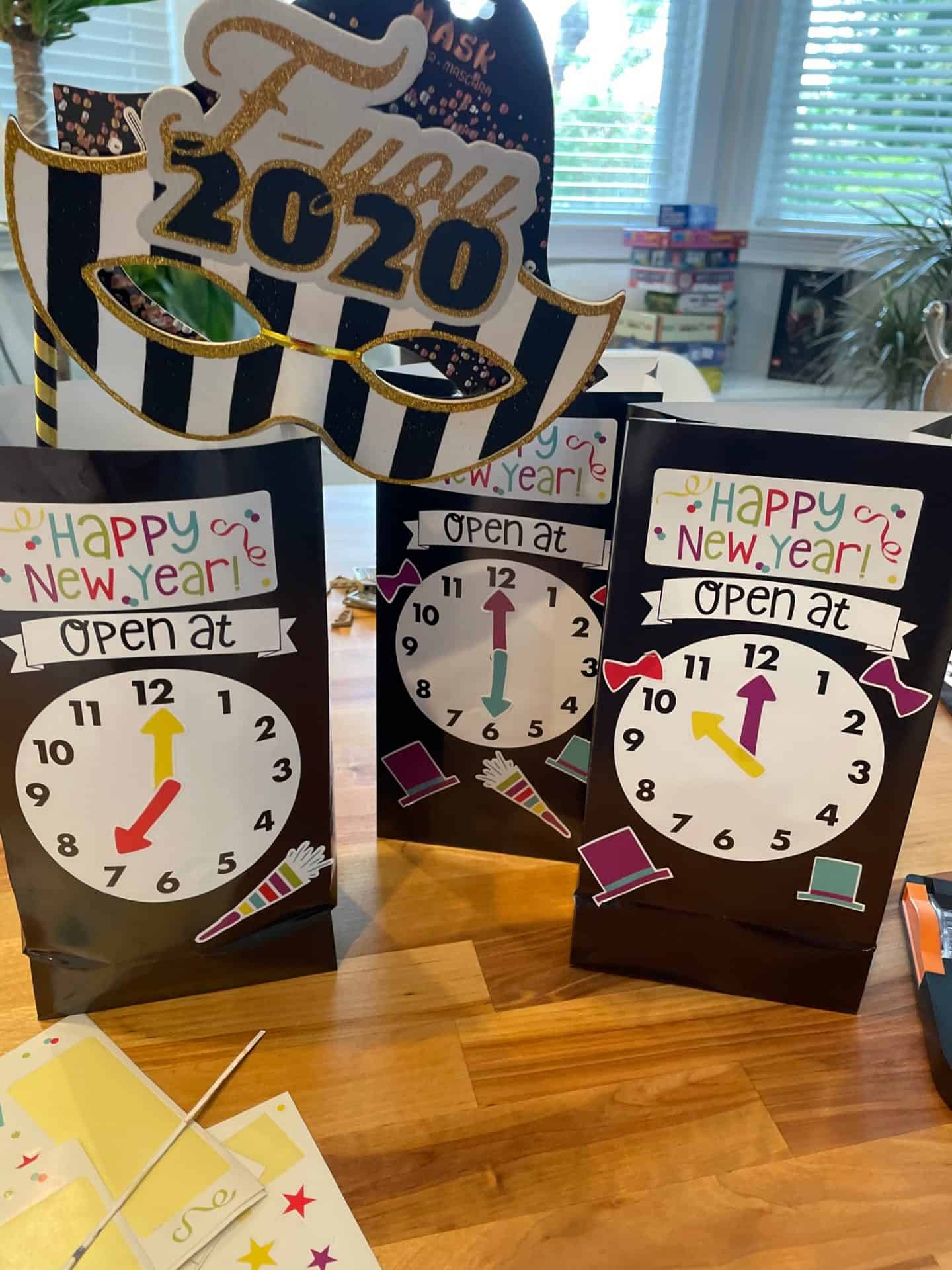 Three DIY New Year's Eve countdown bags decorated with colorful clocks showing different times, labeled “Open at” with festive hats, bows, and noisemakers. Behind them is a black and white striped masquerade mask reading “F-you 2020” in gold glitter. A board game stack and windows are visible in the background.
