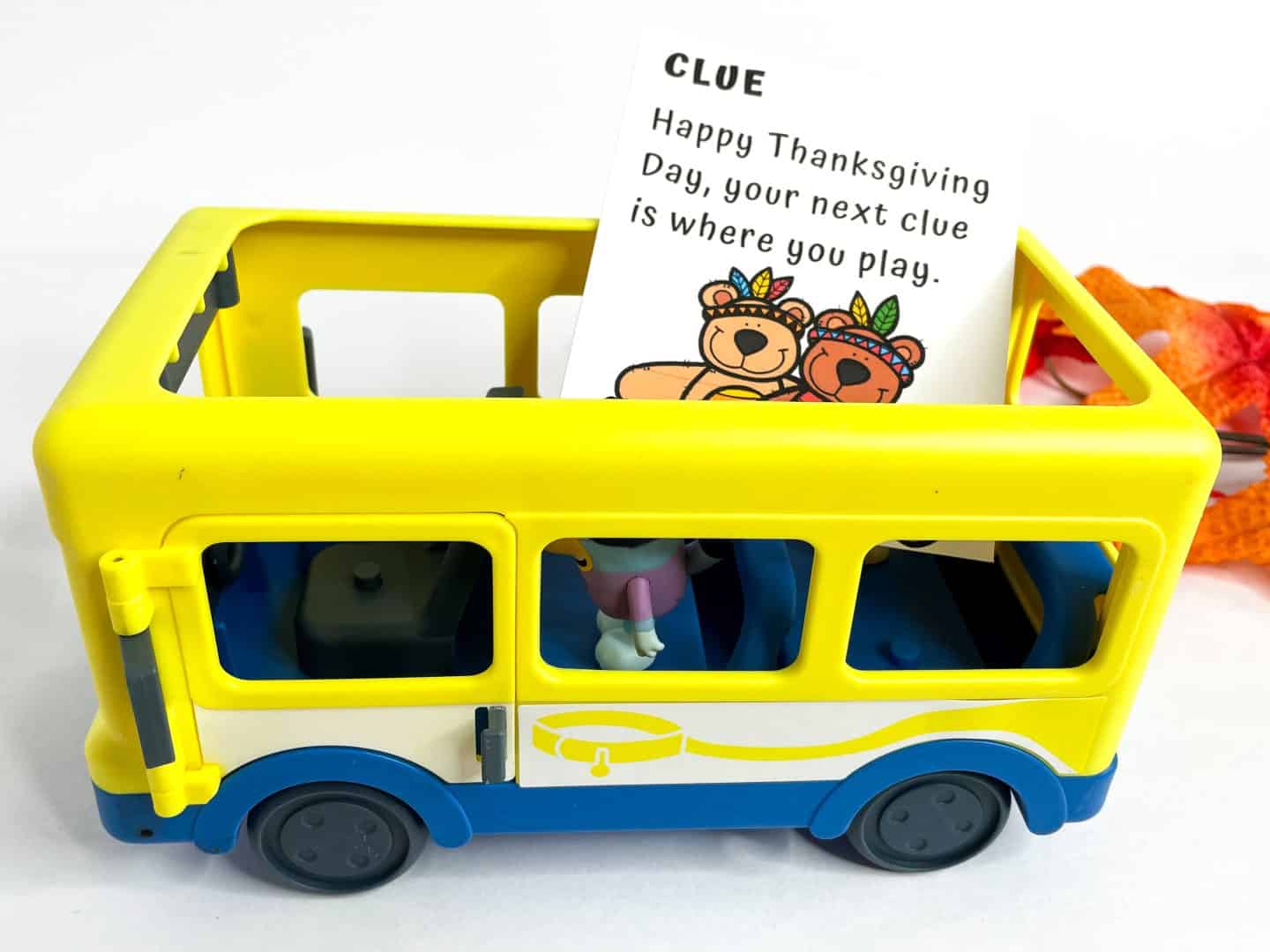 A yellow and blue toy school bus with open top is shown with a Thanksgiving scavenger hunt clue card placed inside. The card reads: “CLUE: Happy Thanksgiving Day, your next clue is where you play,” and features two cartoon bears dressed in festive Thanksgiving attire.