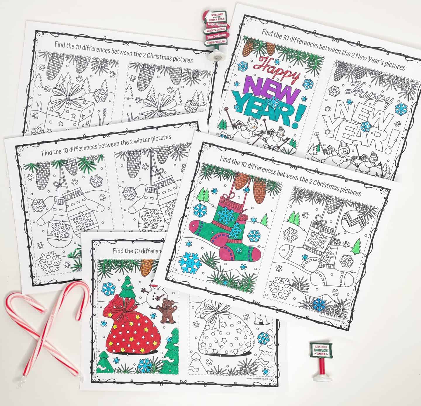 A flat lay of winter-themed “spot the difference” printable activity sheets, each featuring pairs of festive illustrations such as stockings, snowmen, mittens, gifts, and a “Happy New Year” message. Some pages are partially colored in vibrant shades of red, green, purple, and blue. The scene is decorated with candy canes and miniature holiday signs, creating a fun and festive learning vibe.
