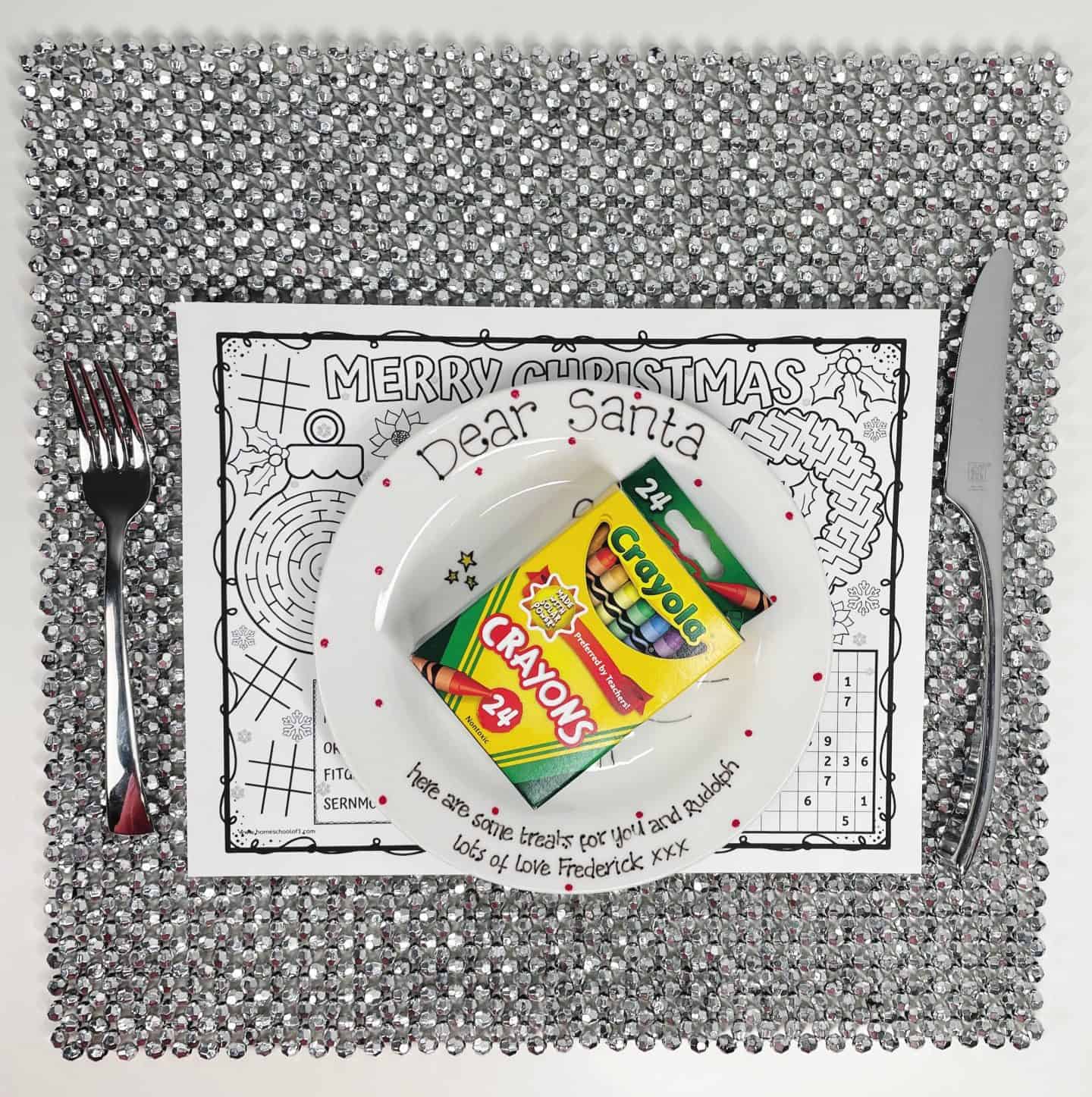 A black and white Christmas activity placemat sits on a sparkly silver mat, featuring festive puzzles and holiday illustrations. On top, a white “Dear Santa” plate holds a box of 24 Crayola crayons, ready for coloring. A fork and knife are placed on either side, mimicking a dining setup.