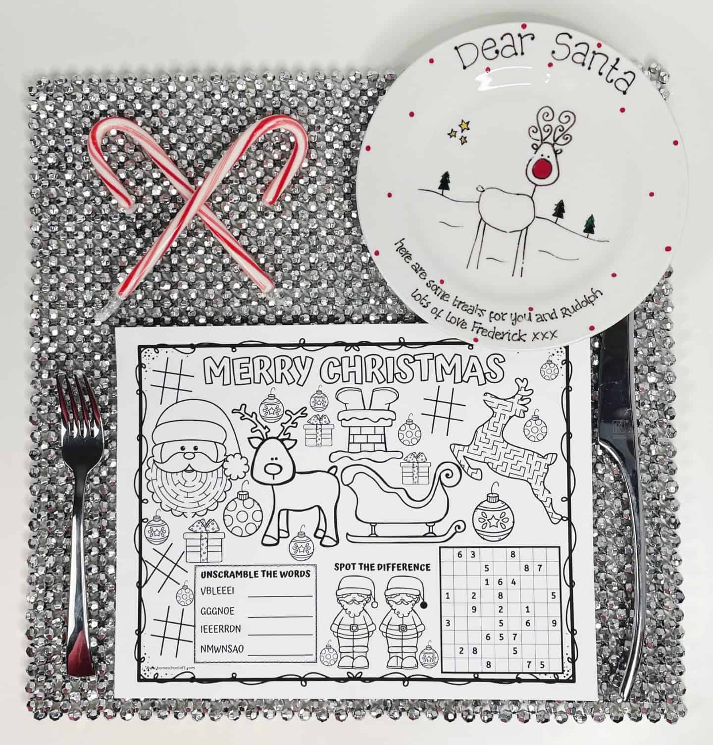 Black and white Christmas activity placemat featuring Santa, reindeer, presents, and games like word unscrambles and spot-the-difference, placed on a silver sequin placemat with a fork and knife. A plate decorated with a reindeer and the words “Dear Santa” sits at the top, with two candy canes crossed beside it.

