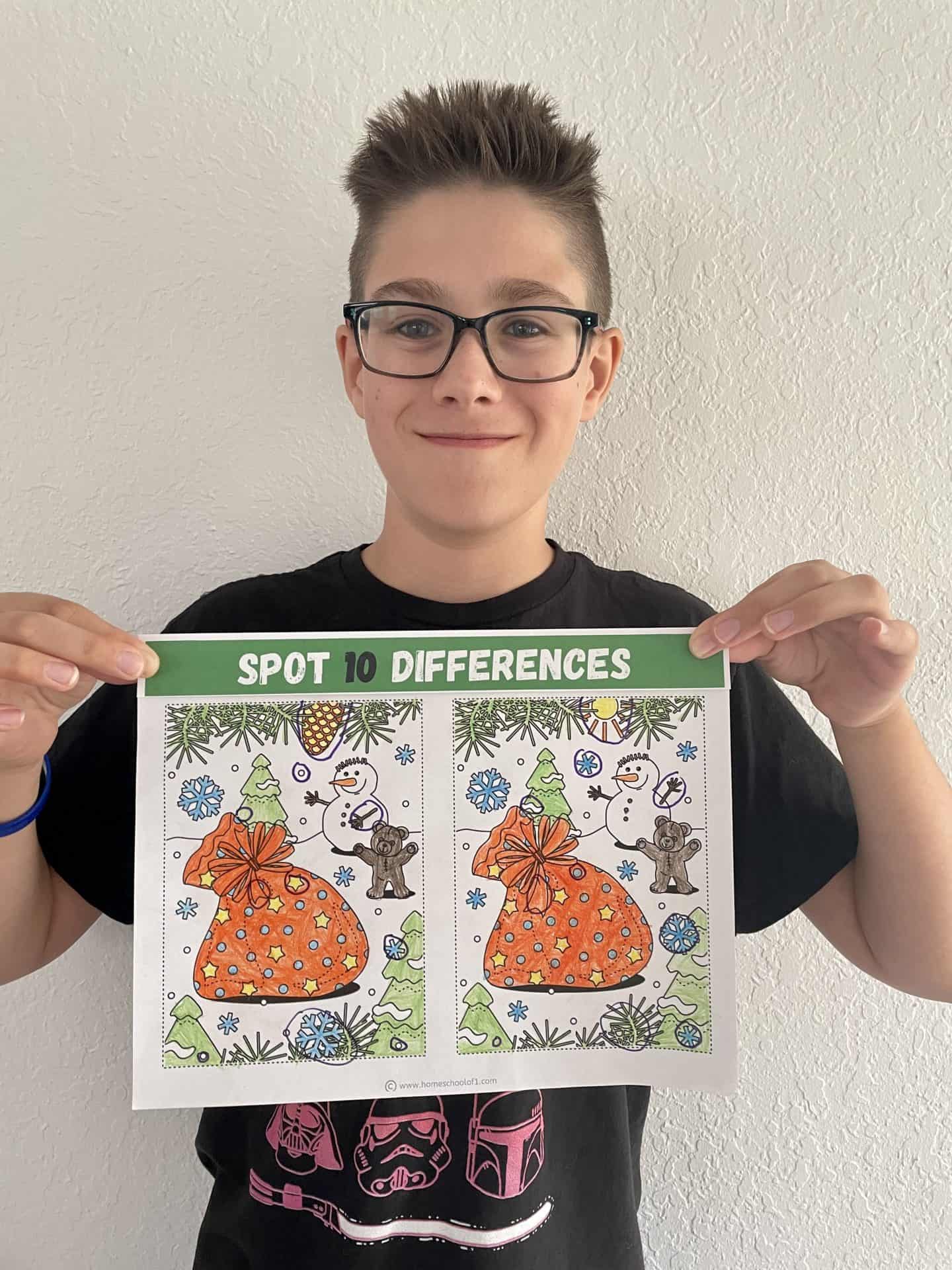 Smiling boy with short brown hair and glasses holds up a completed “Spot 10 Differences” Christmas-themed coloring page. The page features two festive scenes with a large orange gift sack, snowflakes, a snowman, trees, and a teddy bear, all fully colored. He wears a black Star Wars shirt, and the background is a plain white wall.