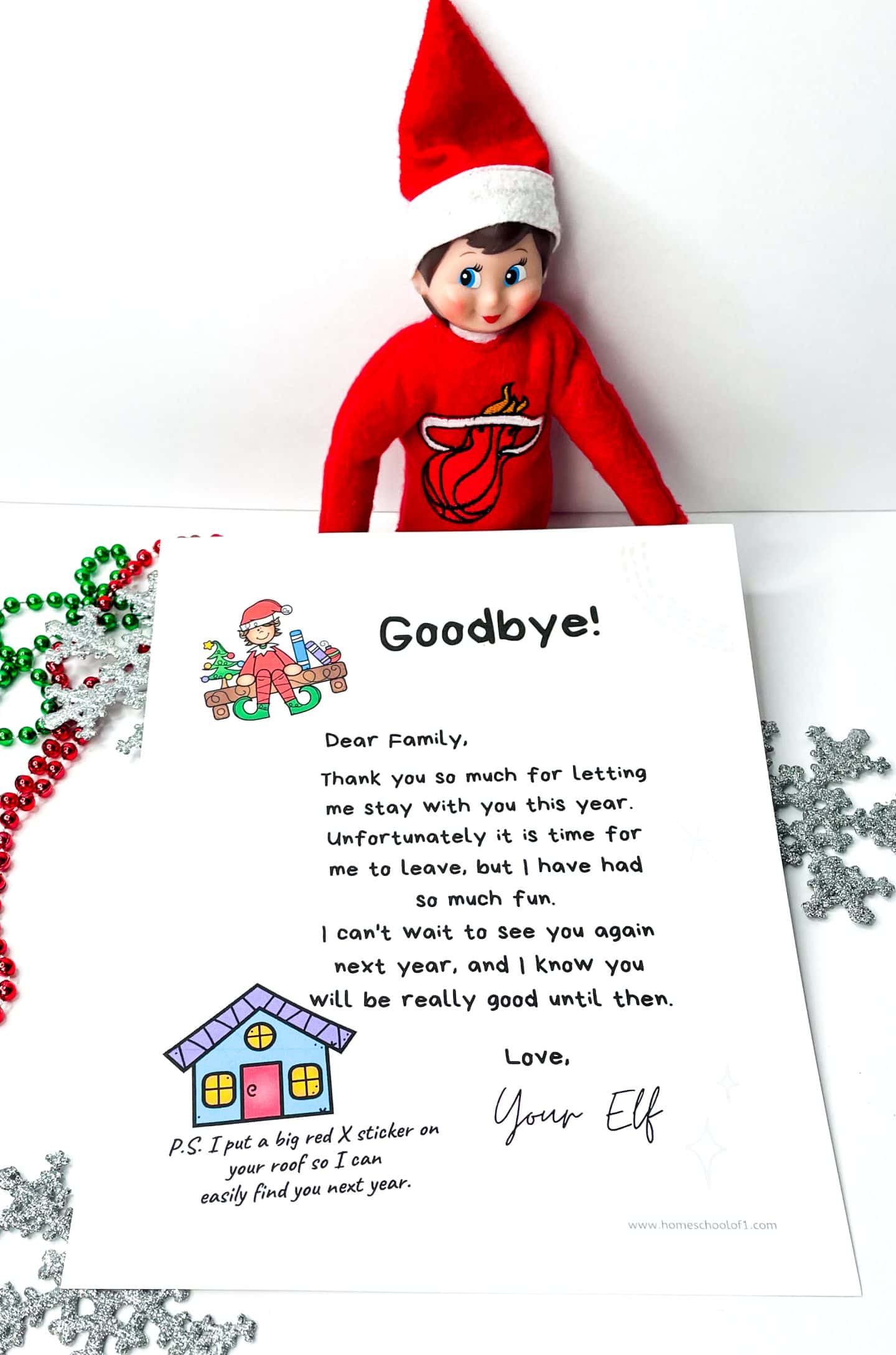 An Elf on the Shelf doll dressed in a red Miami Heat sweater and Santa hat stands behind a printed farewell letter. The letter, titled "Goodbye!", thanks the family for a fun time, mentions the elf must leave, and promises to return next year. It ends with a playful note saying a red X was placed on the roof so the elf can easily find the house again. Decorative red and green beads and glittery silver snowflakes surround the setup.

