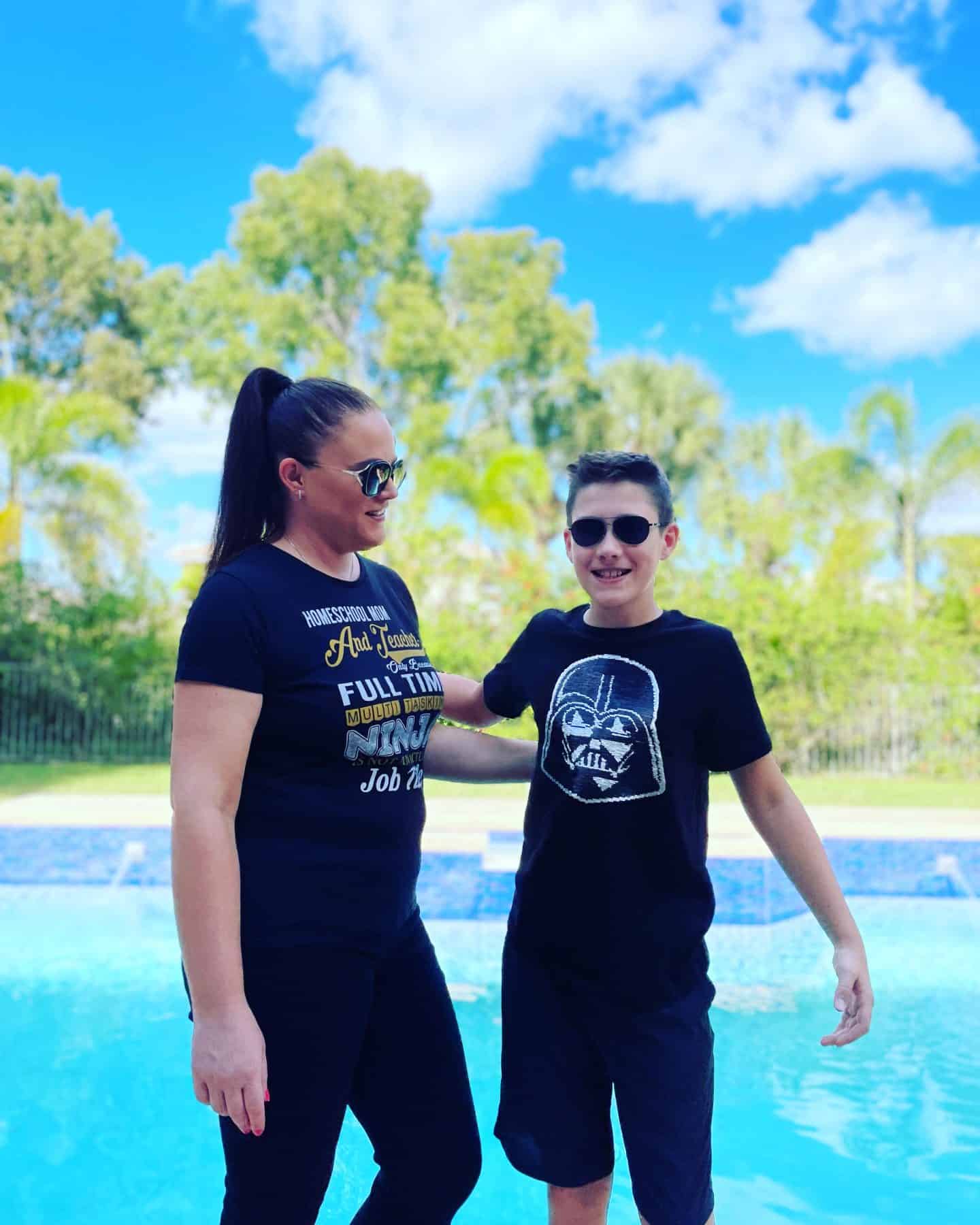 Clare Brown, founder of Homeschool of 1, smiling beside her son by the pool, both wearing black shirts and sunglasses, with palm trees in the background.