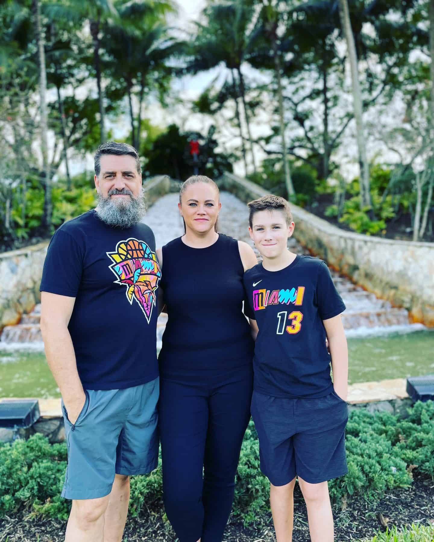 Clare Brown of Homeschool of 1 standing with her husband and son in front of a tropical garden path with palm trees and a stone waterfall. All three are wearing Miami-themed shirts.