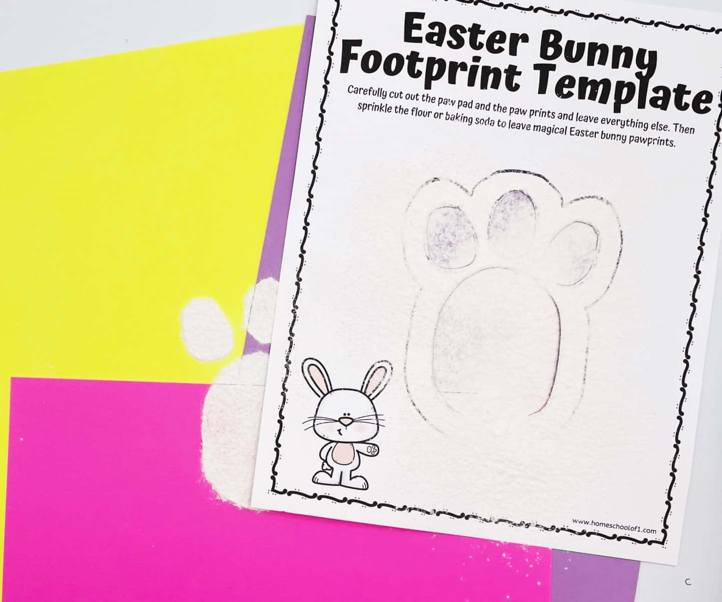 An Easter Bunny Footprint Template with cut-out paw shapes lies on top of a vibrant pink, purple, and yellow paper background, dusted with flour or baking soda. A completed white bunny pawprint is visible on the surface underneath, showing how the stencil was used to create the magical footprint. A small cartoon bunny decorates the lower corner of the template, adding a playful touch.


