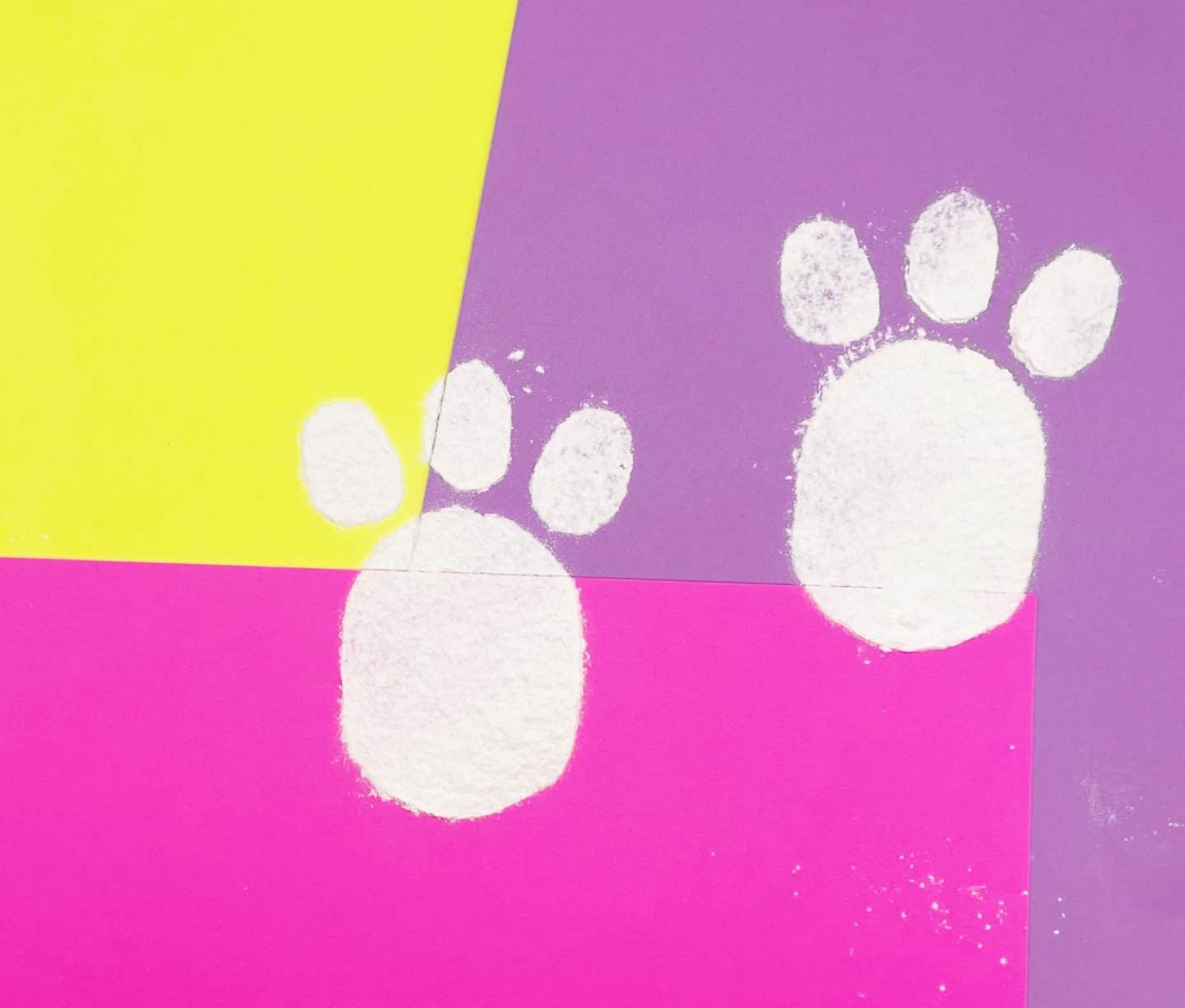 Two white Easter bunny footprints made with flour or baking soda are stamped onto a colorful background of pink, yellow, and purple paper. Each footprint features a large oval pad with three smaller toe shapes above, resembling a hopping trail left by the Easter Bunny. The bright, playful setting creates a magical Easter morning vibe.