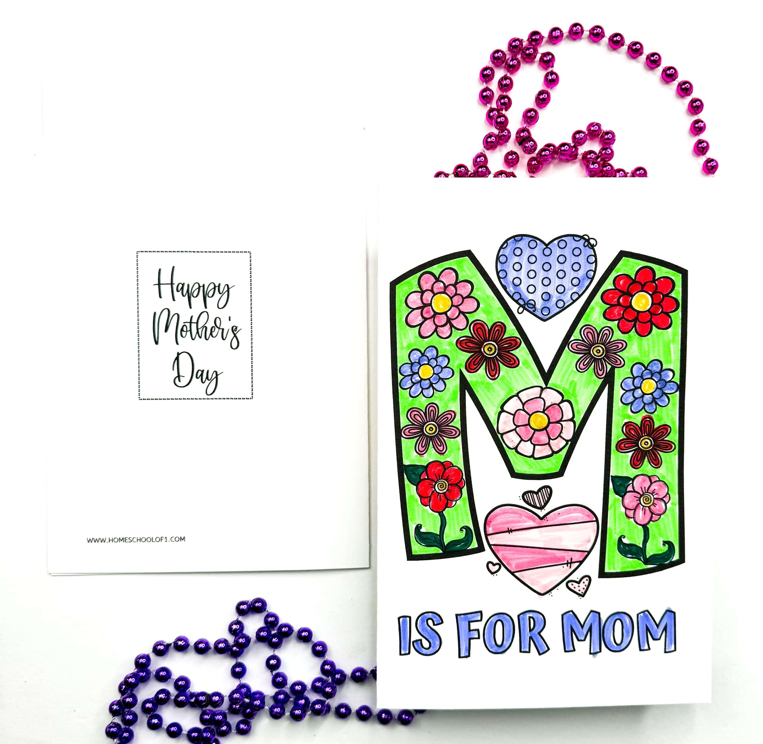Colorful homemade Mother’s Day card with a large, flower-filled letter "M" colored in green, red, blue, pink, and purple. Below it, bold text reads “IS FOR MOM,” and the inside of the card says “Happy Mother’s Day” in cursive font. Purple and pink beaded necklaces are scattered around for decoration.

