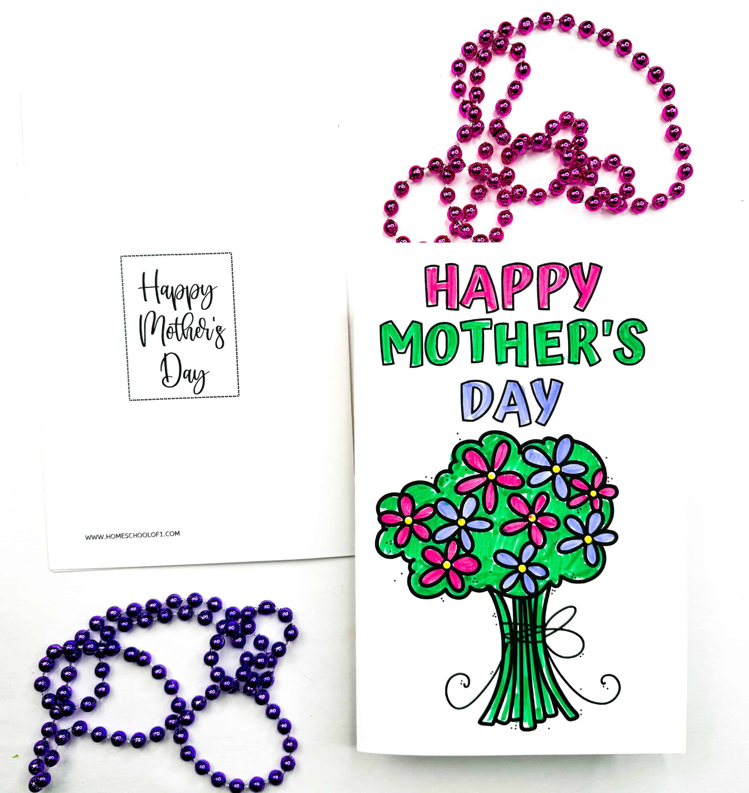 Handmade Mother's Day card featuring a colorful bouquet of pink and purple flowers with green stems, tied with a bow. Above the bouquet, the card reads “HAPPY MOTHER’S DAY” in bold, multi-colored letters. A second card in the background shows a printed “Happy Mother’s Day” message in cursive, and the scene is decorated with pink and purple beaded necklaces.