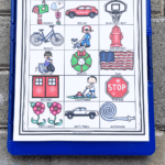 Colorful neighborhood scavenger hunt printable clipped to a blue clipboard, featuring 15 illustrated items for kids to find outdoors including a red car, basketball hoop, dog, trash can, lawn mower, and stop sign. Each item has a matching word label, encouraging kids to explore their surroundings while building observation skills.