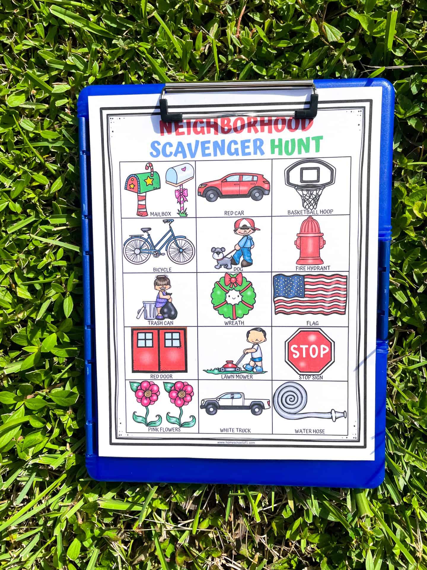 Colorful neighborhood scavenger hunt printable on a blue clipboard resting on green grass. The activity sheet includes illustrated items like a red car, basketball hoop, dog, fire hydrant, wreath, lawn mower, and pink flowers for kids to find outdoors.