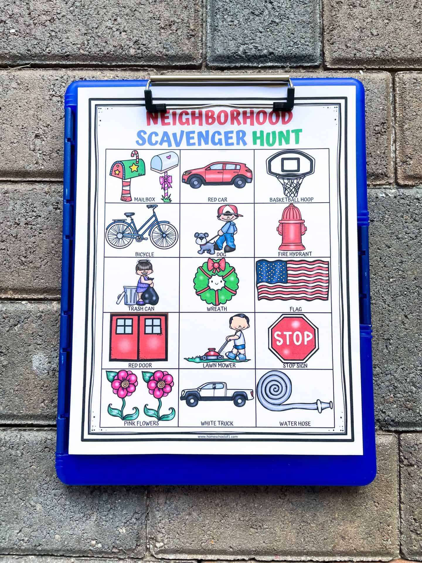 Neighborhood scavenger hunt printable clipped to a blue clipboard on a brick surface. The worksheet includes colorful illustrations of items to find like a mailbox, red car, basketball hoop, dog, trash can, flag, lawn mower, and stop sign.