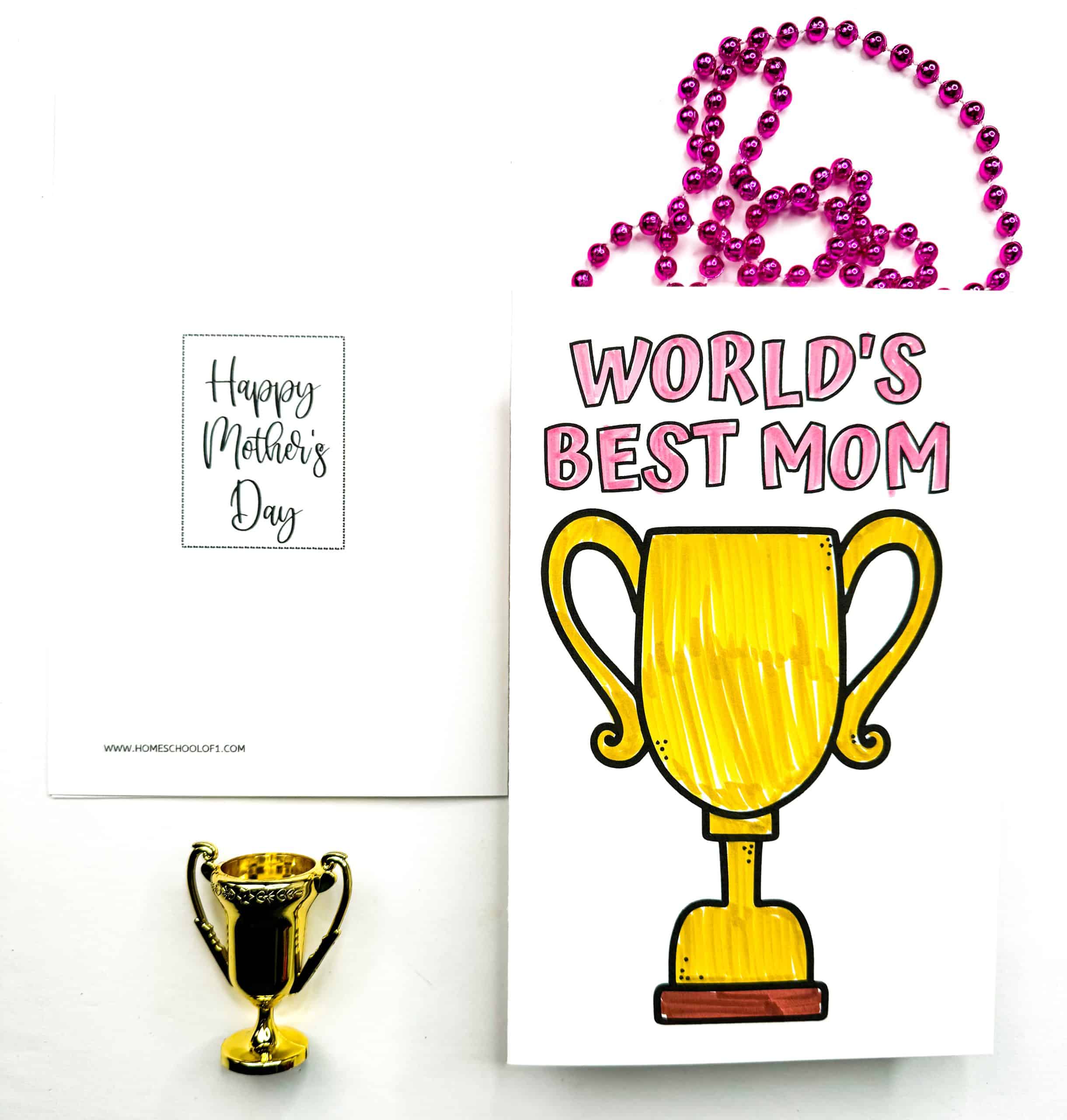 Mother’s Day card featuring a large, hand-colored golden trophy and the bold message “WORLD’S BEST MOM” in pink letters above it. A small gold trophy sits beside the card, and the background includes a second card with “Happy Mother’s Day” in cursive and a strand of shiny pink beads.