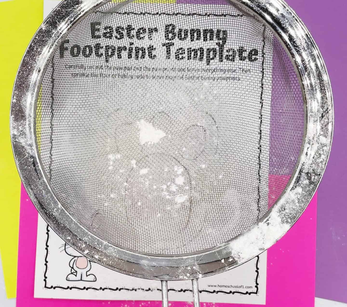 A metal flour sifter is held over an Easter Bunny Footprint Template placed on colorful pink, yellow, and purple paper. White powder, likely flour or baking soda, has been sprinkled through the sifter onto the stencil, creating a bunny pawprint shape. The setup demonstrates how to use the stencil to make magical Easter bunny tracks as part of a fun holiday activity.

