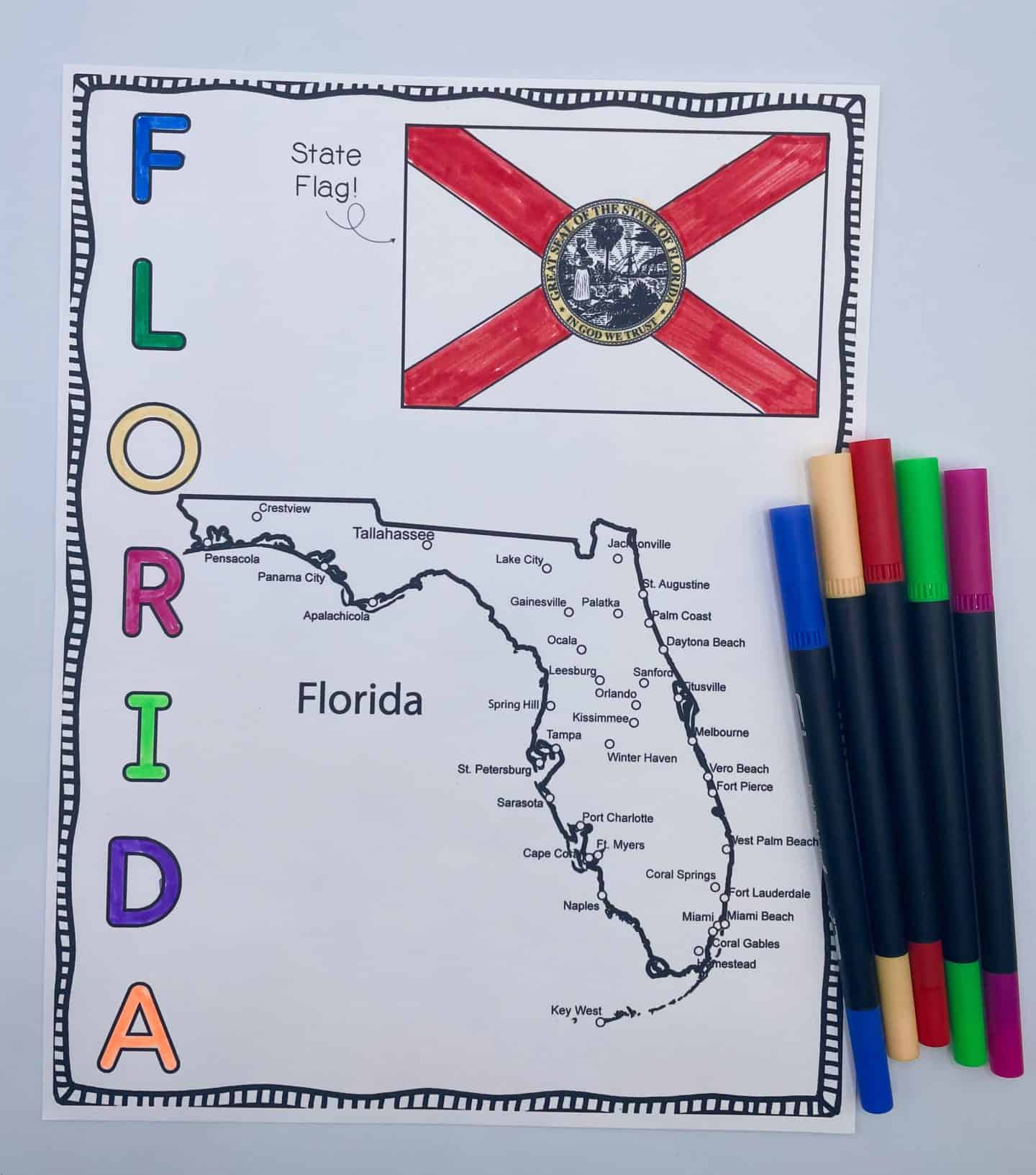 A Florida state coloring page for kids, featuring a black-and-white map labeled with major cities and a fully colored state flag with a red X and state seal. The word “FLORIDA” is written vertically in rainbow-colored block letters. Several colorful markers are placed to the right of the page.

