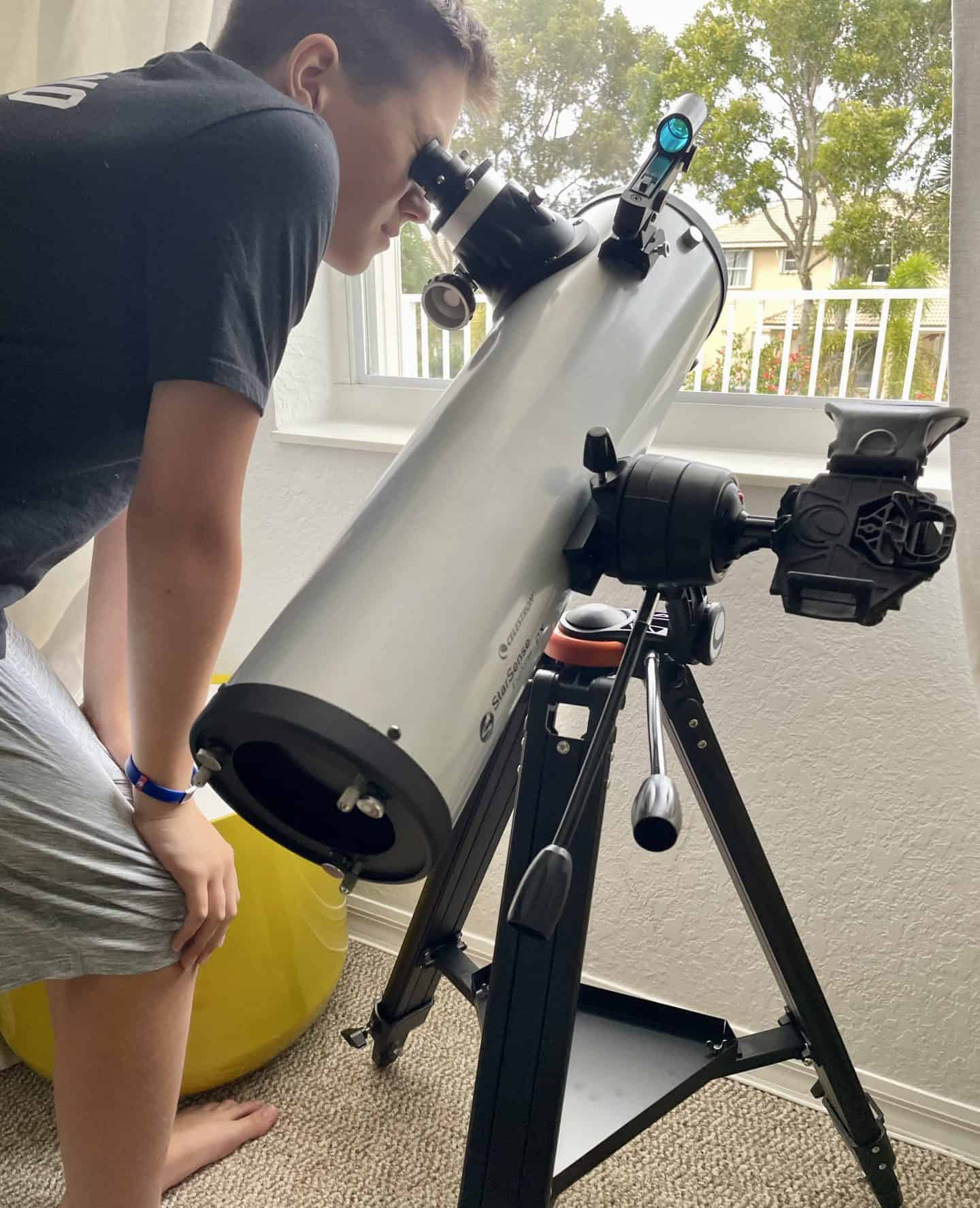 A young person peers through the eyepiece of a large silver telescope set up indoors near a window. The telescope is mounted on a black tripod stand, and there's a smartphone holder attached for capturing images. Outside the window, trees and part of a house are visible, suggesting a daytime observation or setup.