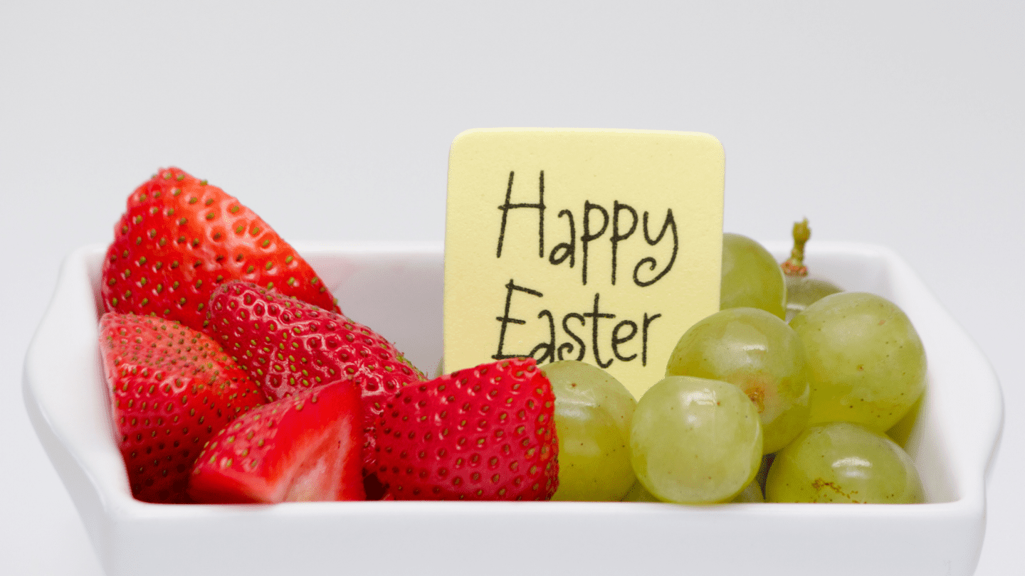 Close-up of a white dish filled with fresh strawberries and green grapes, with a small yellow sign tucked in the middle that reads “Happy Easter” in playful handwriting. The clean background and vibrant fruit give a healthy, cheerful holiday vibe.