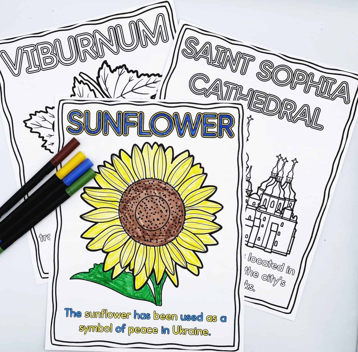 Three Ukraine-themed coloring pages are shown, with the main focus on a completed “Sunflower” page featuring a bright yellow sunflower with a brown center and green leaves. The text below reads, “The sunflower has been used as a symbol of peace in Ukraine.” Behind it are two partially visible pages titled “Viburnum” and “Saint Sophia Cathedral.” A few markers in green, blue, yellow, brown, and black are placed to the left.