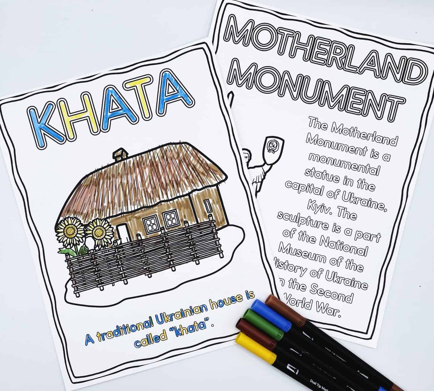 Two Ukraine-themed coloring pages. The front page is titled “Khata” and shows a traditional Ukrainian house with a thatched roof, a wooden fence, and sunflowers in front. The word “Khata” is colored in blue and yellow, and the text below reads, “A traditional Ukrainian house is called ‘khata’.” Behind it is a page titled “Motherland Monument,” partially colored, with descriptive text about the monument in Kyiv. Colored markers are placed beside the pages.