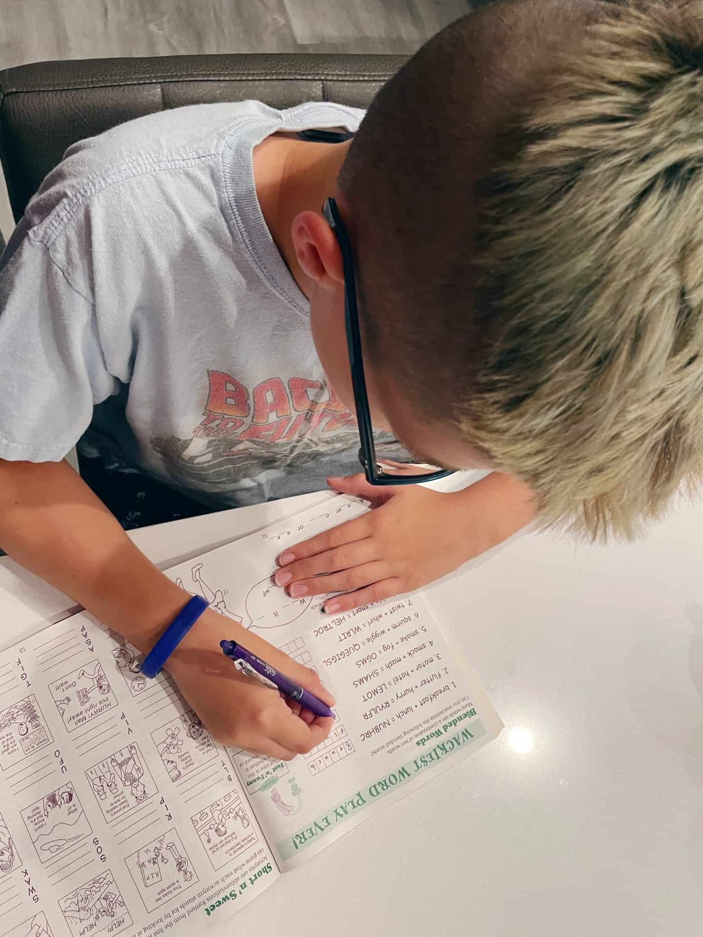 A child with short blond and brown hair, wearing glasses and a “Back to the Future” T-shirt, works on a word puzzle in a printed activity book. The page is titled “Wackiest Word Play Ever!” and includes word scrambles, a word wheel, and cartoon-style illustrations.