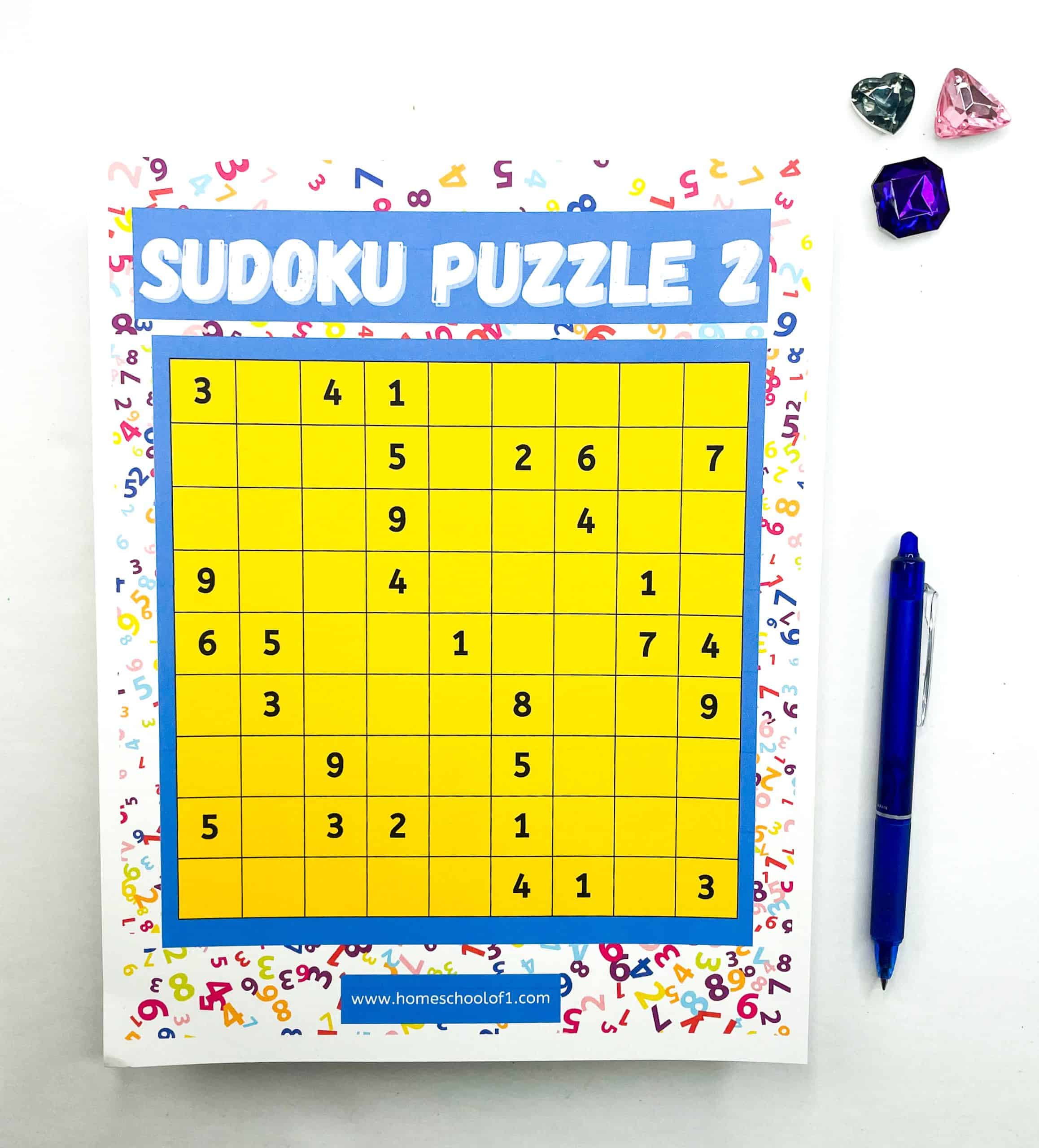 A bright yellow Sudoku grid labeled “Sudoku Puzzle 2” is partially filled in with black numbers and bordered by a white frame decorated with colorful, scattered numerals. A blue pen lies to the right of the puzzle, next to four gem-like decorations in pink, purple, and clear heart and jewel shapes. 