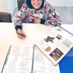 Middle school student sitting at a table working on a History Odyssey lesson, with the curriculum binder and a history reference book open in front of him.