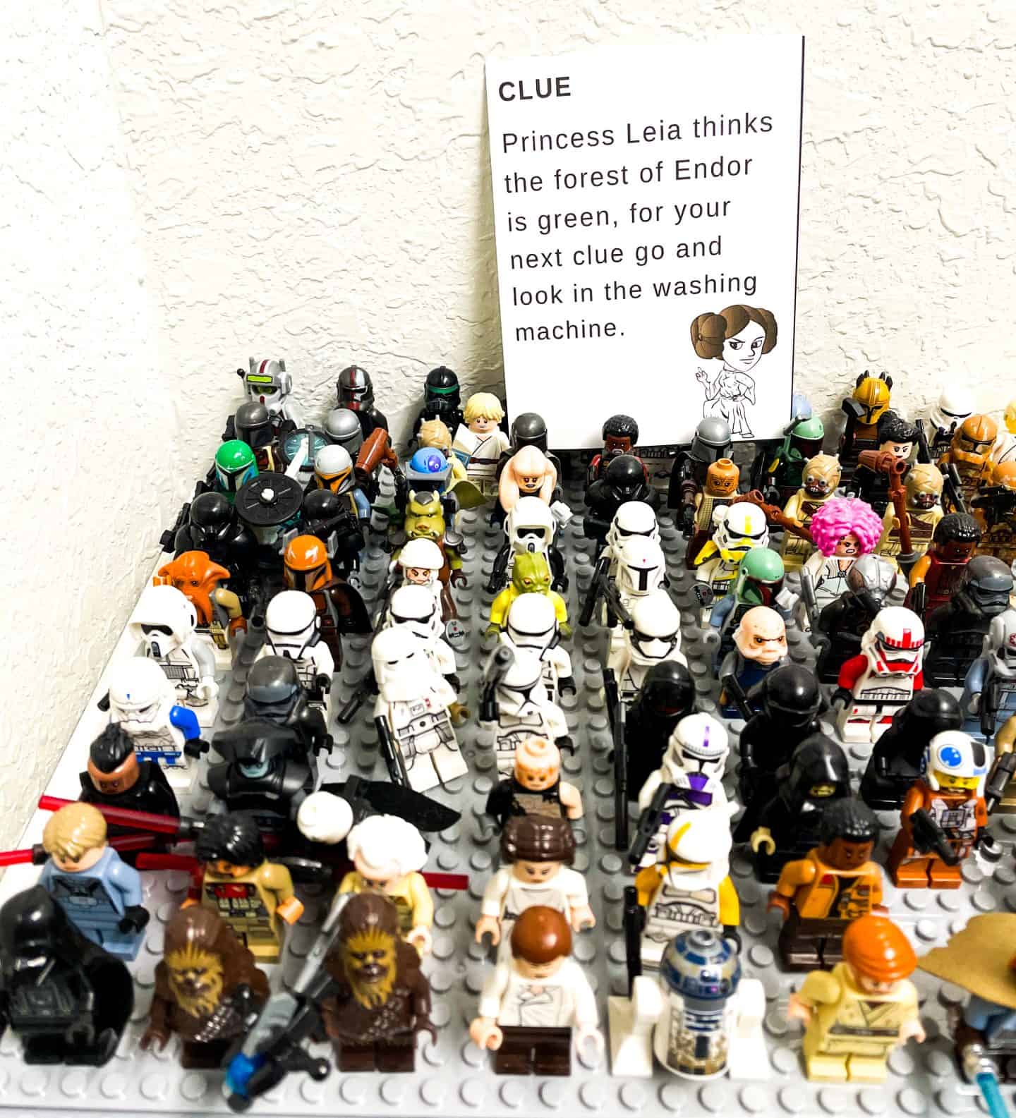 A Star Wars scavenger hunt clue card featuring Princess Leia is placed behind a large display of Star Wars LEGO minifigures, including stormtroopers, Chewbacca, and R2D2. The clue reads, “Princess Leia thinks the forest of Endor is green, for your next clue go and look in the washing machine.”

