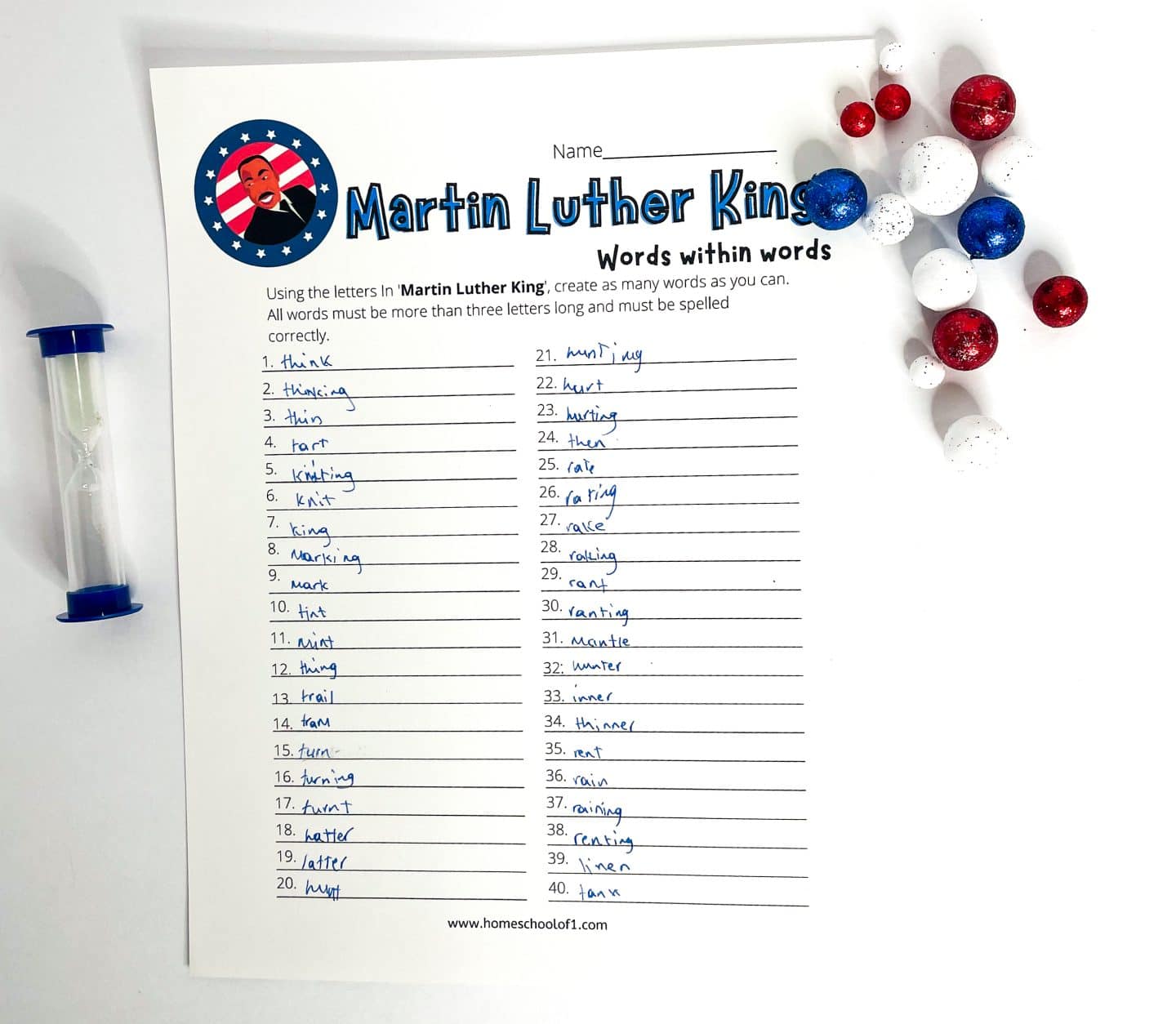 A “Words Within Words” worksheet themed around Martin Luther King Jr. is filled with 40 handwritten words made from the letters in “Martin Luther King.” The page features a cartoon-style patriotic illustration of MLK in a circular emblem, and red, white, and blue decorative spheres rest on the right side. A blue sand timer sits to the left, suggesting a timed word game challenge.