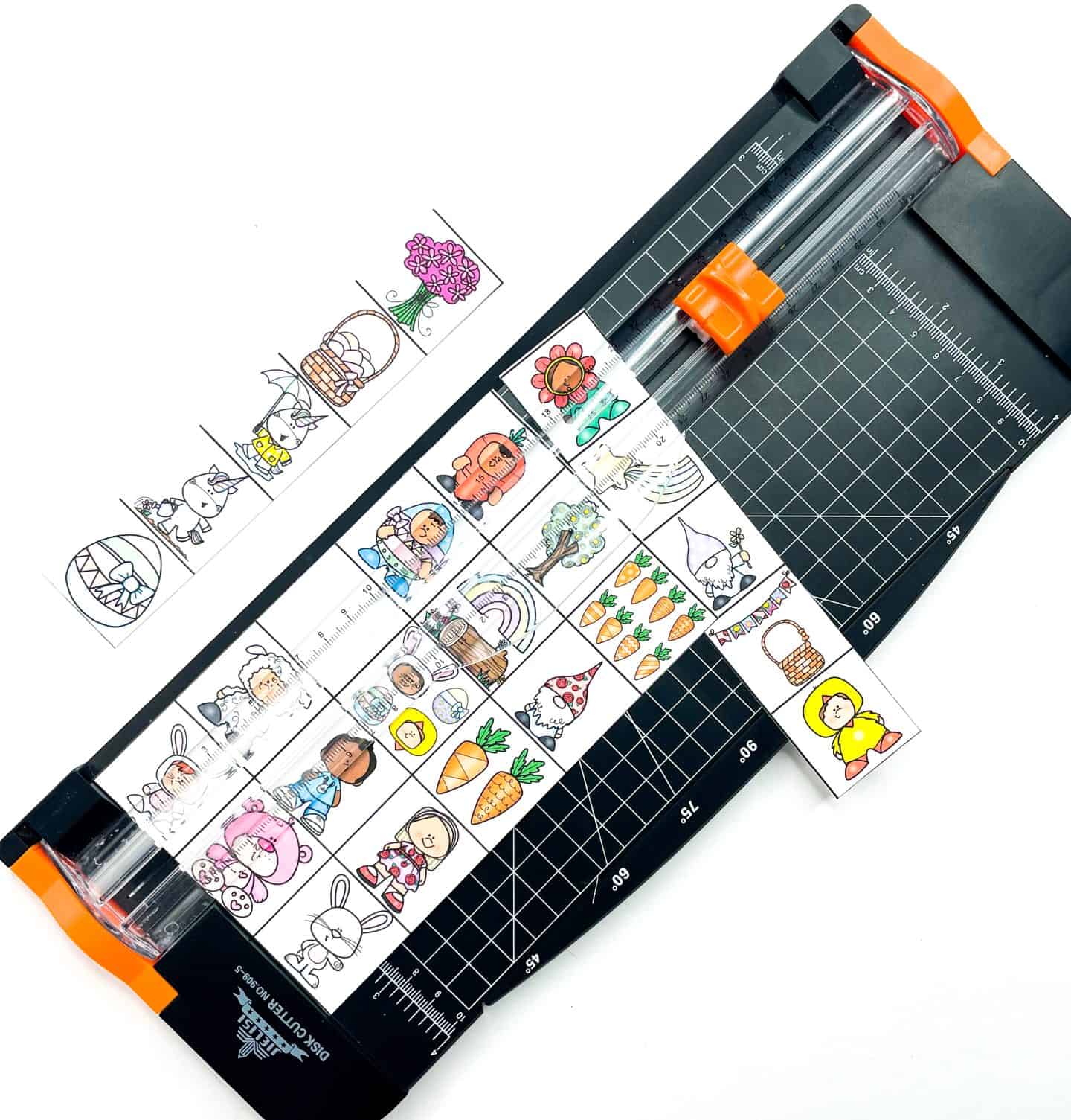 Easter-themed bingo game pieces being cut with a paper trimmer, featuring colorful illustrations of bunnies, chicks, Easter baskets, gnomes, carrots, flowers, and more. The small square images are arranged in rows, ready to be used as calling cards or game pieces. The setup is part of a DIY printable Easter bingo game