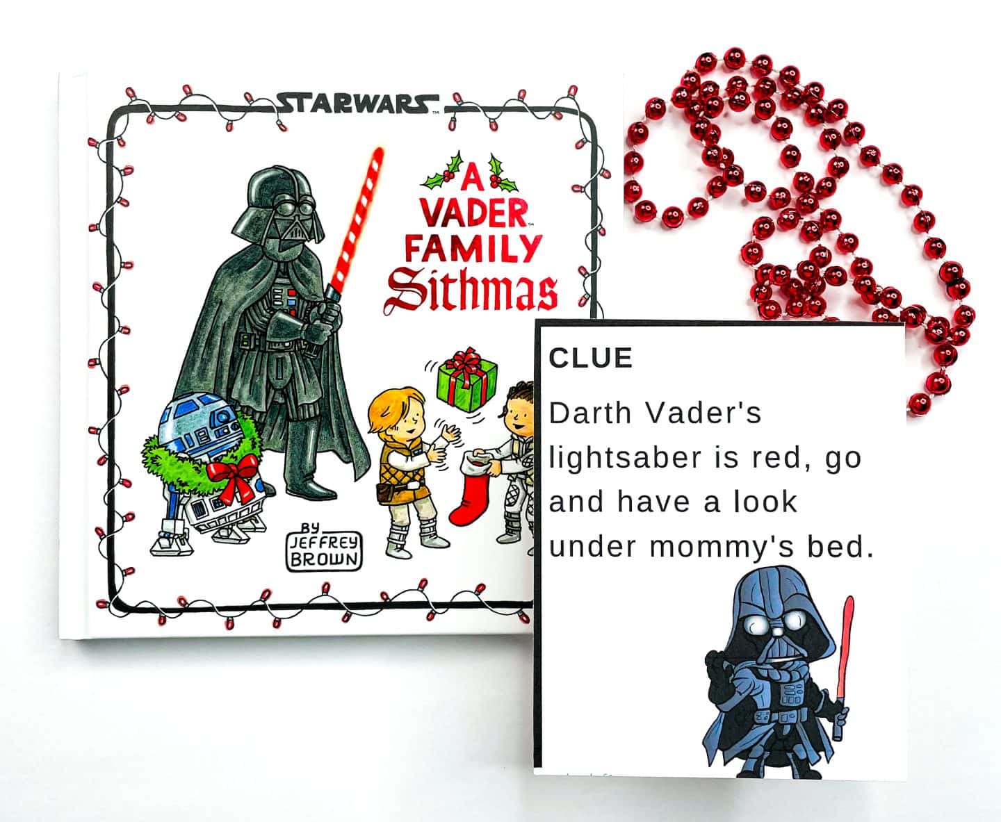 A "Star Wars scavenger hunt clues" card featuring Darth Vader is placed beside the holiday-themed book A Vader Family Sithmas by Jeffrey Brown. The clue reads, “Darth Vader's lightsaber is red, go and have a look under mommy's bed,” with red holiday beads completing the festive look.

