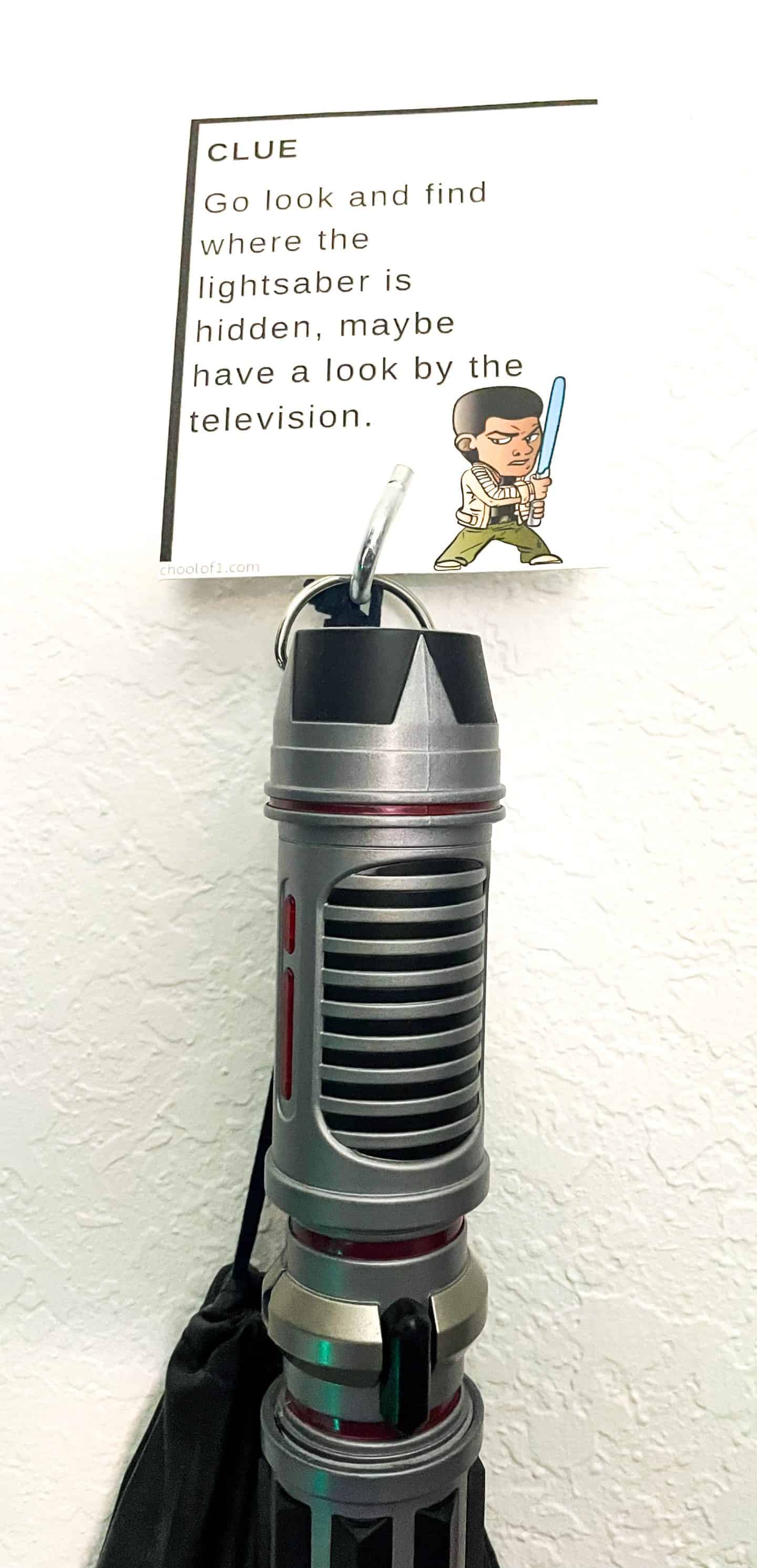 A Star Wars scavenger hunt clue card featuring Finn is displayed above a toy lightsaber. The clue reads, “Go look and find where the lightsaber is hidden, maybe have a look by the television,” hinting at the next step in the galactic-themed game.

