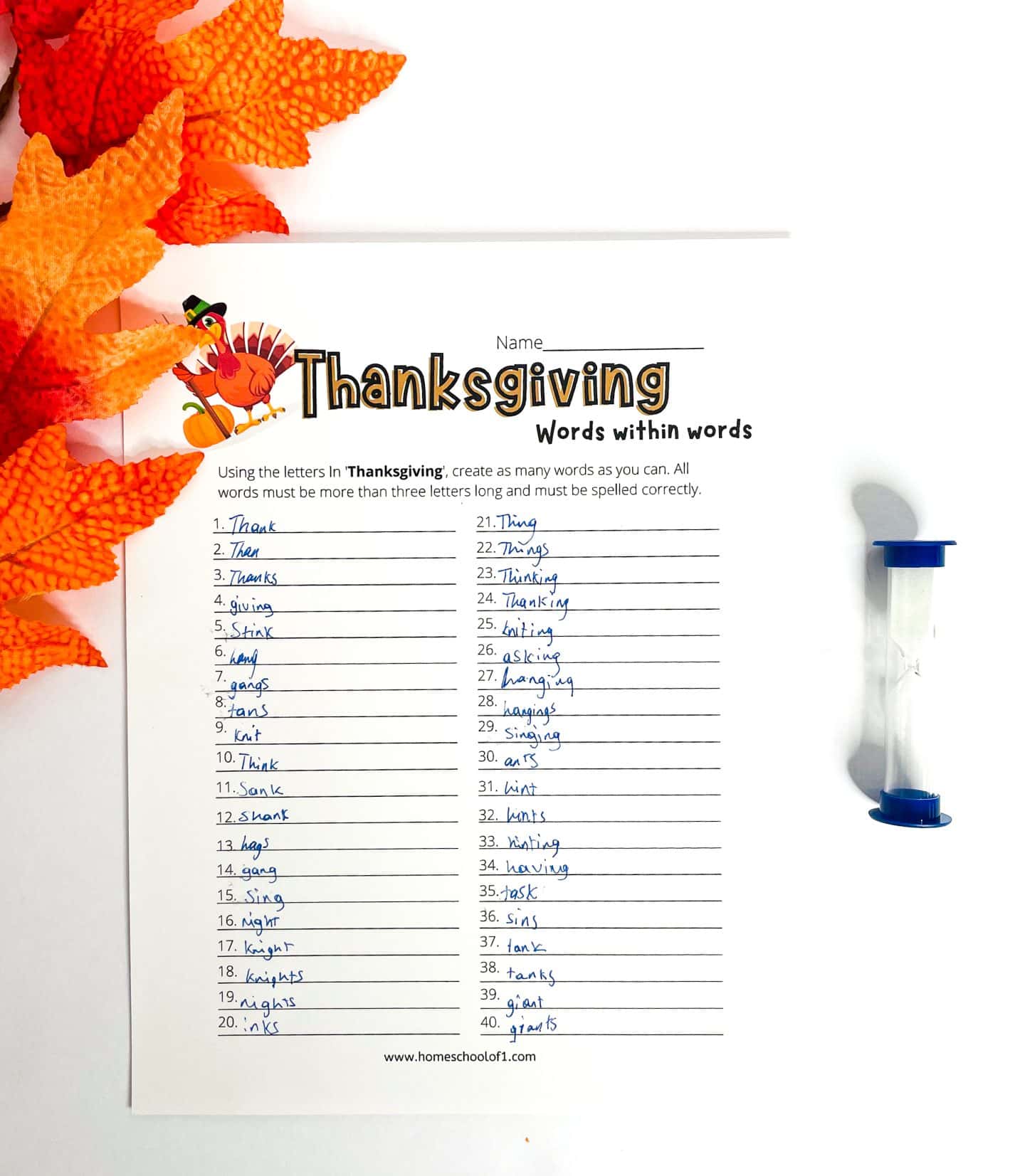A Thanksgiving-themed “Words Within Words” worksheet is shown completed with 40 handwritten words using letters from the word “Thanksgiving.” The page features a cartoon turkey illustration and instructions to form words longer than three letters. Fall-colored fabric leaves and a blue sand timer are placed beside the sheet, creating a festive and educational atmosphere.