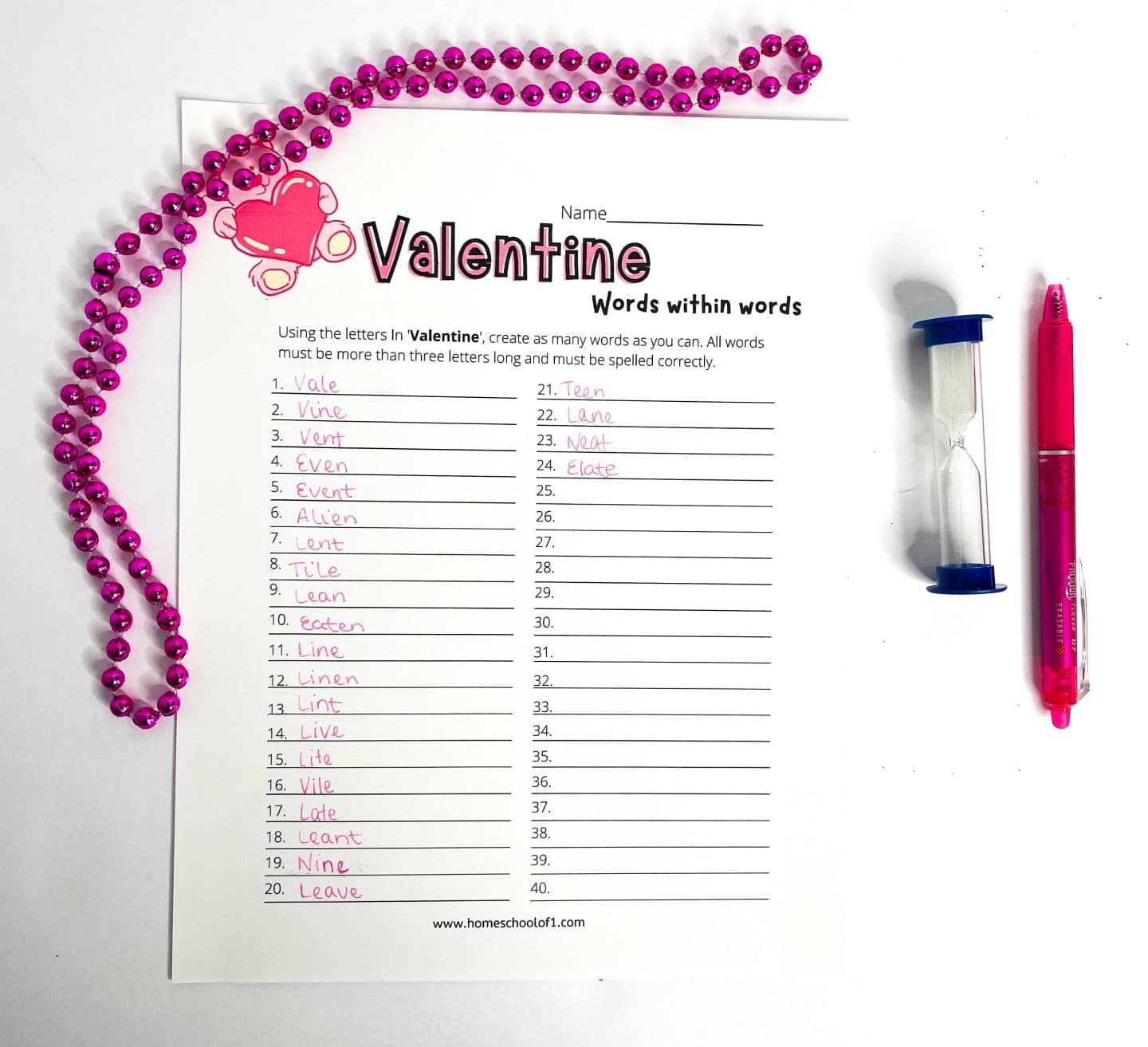 A Valentine's Day-themed “Words Within Words” worksheet is partially filled with 24 handwritten words formed from the letters in "Valentine." The sheet features a pink heart and bell graphic, with pink beaded garland draped on the left, and a pink pen and blue sand timer on the right, creating a festive, love-themed learning setup.