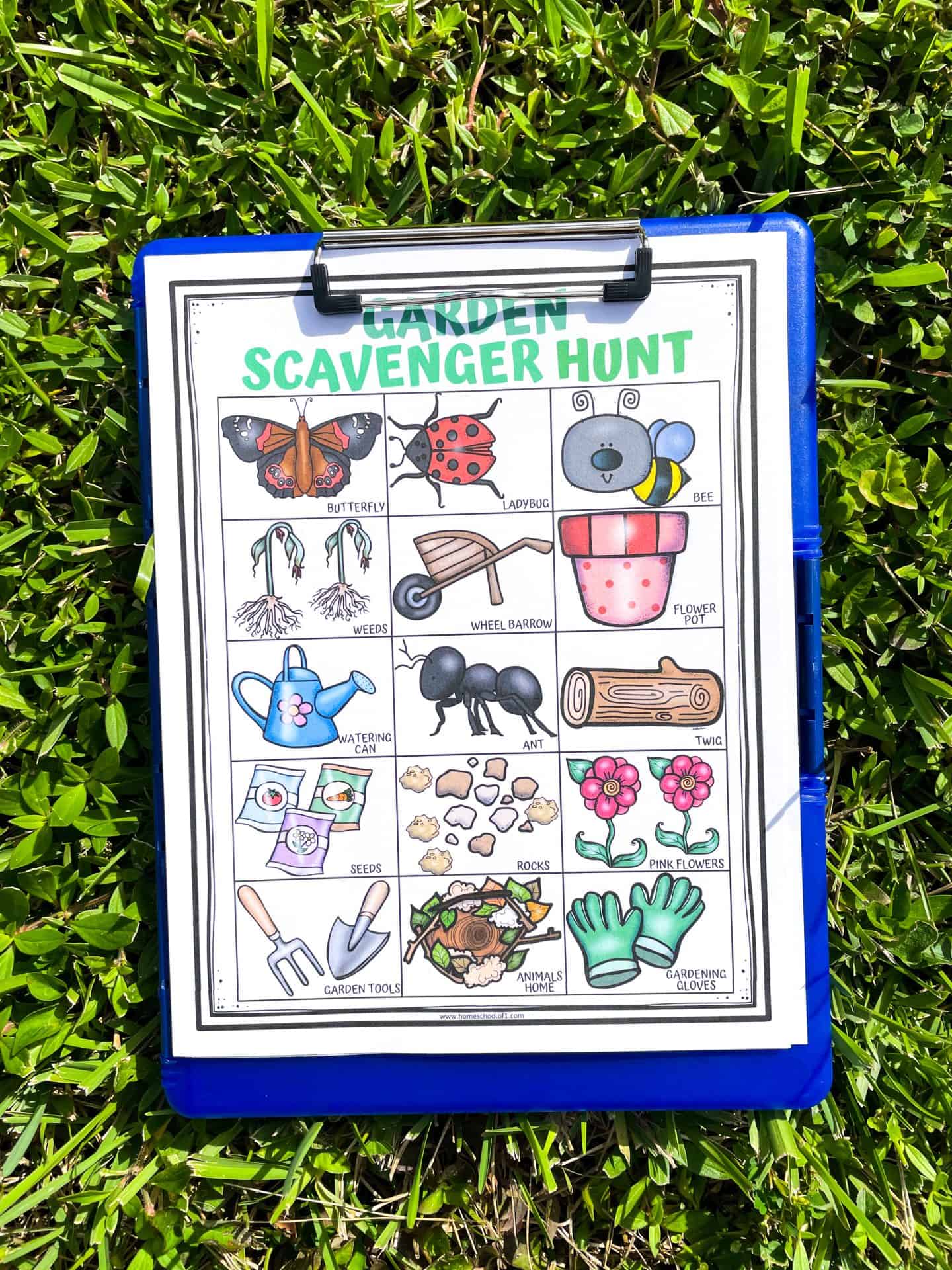 A garden scavenger hunt printable clipped to a blue clipboard resting on green grass. The sheet features colorful illustrations of garden items like a butterfly, ladybug, bee, wheelbarrow, flower pot, ant, watering can, pink flowers, and more, arranged in a 4x4 grid format for kids to find during outdoor play.

