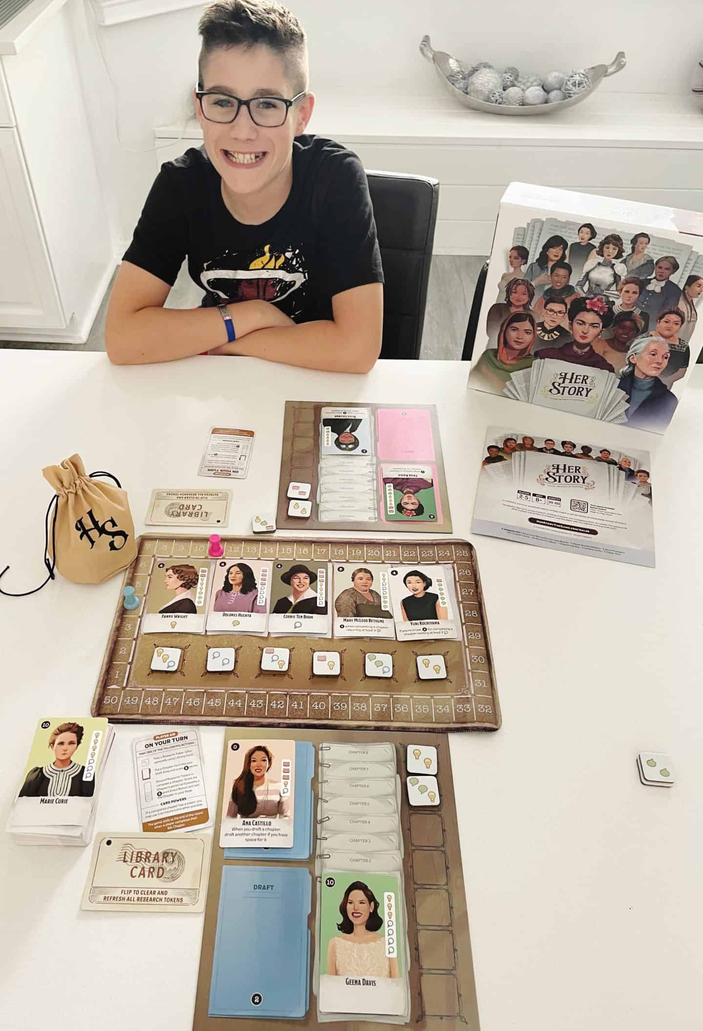 A smiling child sits at a white table playing the board game HerStory, which features influential women from history. The game board is laid out with character cards including Marie Curie, Geena Davis, and Dolores Huerta, along with tokens and a drawstring bag labeled “HS.” The game box stands upright nearby, displaying illustrated portraits of women such as Ruth Bader Ginsburg, Malala Yousafzai, and Frida Kahlo.