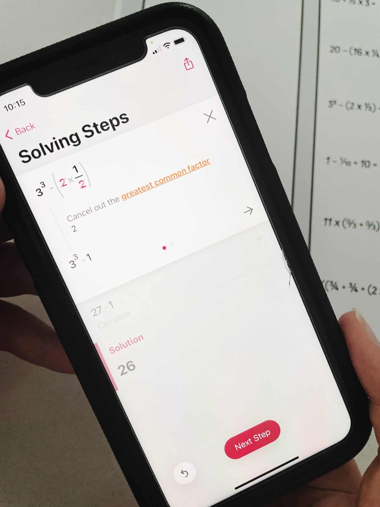 A smartphone screen displaying a math-solving app with the title "Solving Steps." The app highlights canceling out the greatest common factor and shows the solution as 26. A "Next Step" button is visible at the bottom of the screen. A hand holds the phone, and part of a math worksheet is seen in the background.