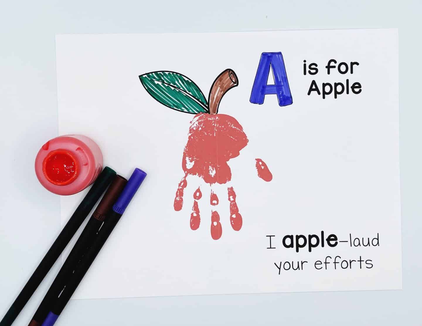 A finished "A is for Apple" handprint craft shows a red handprint shaped like an apple with added green leaves and a brown stem. The phrase "I apple-laud your efforts" is printed underneath, with markers and red paint sitting beside the artwork.