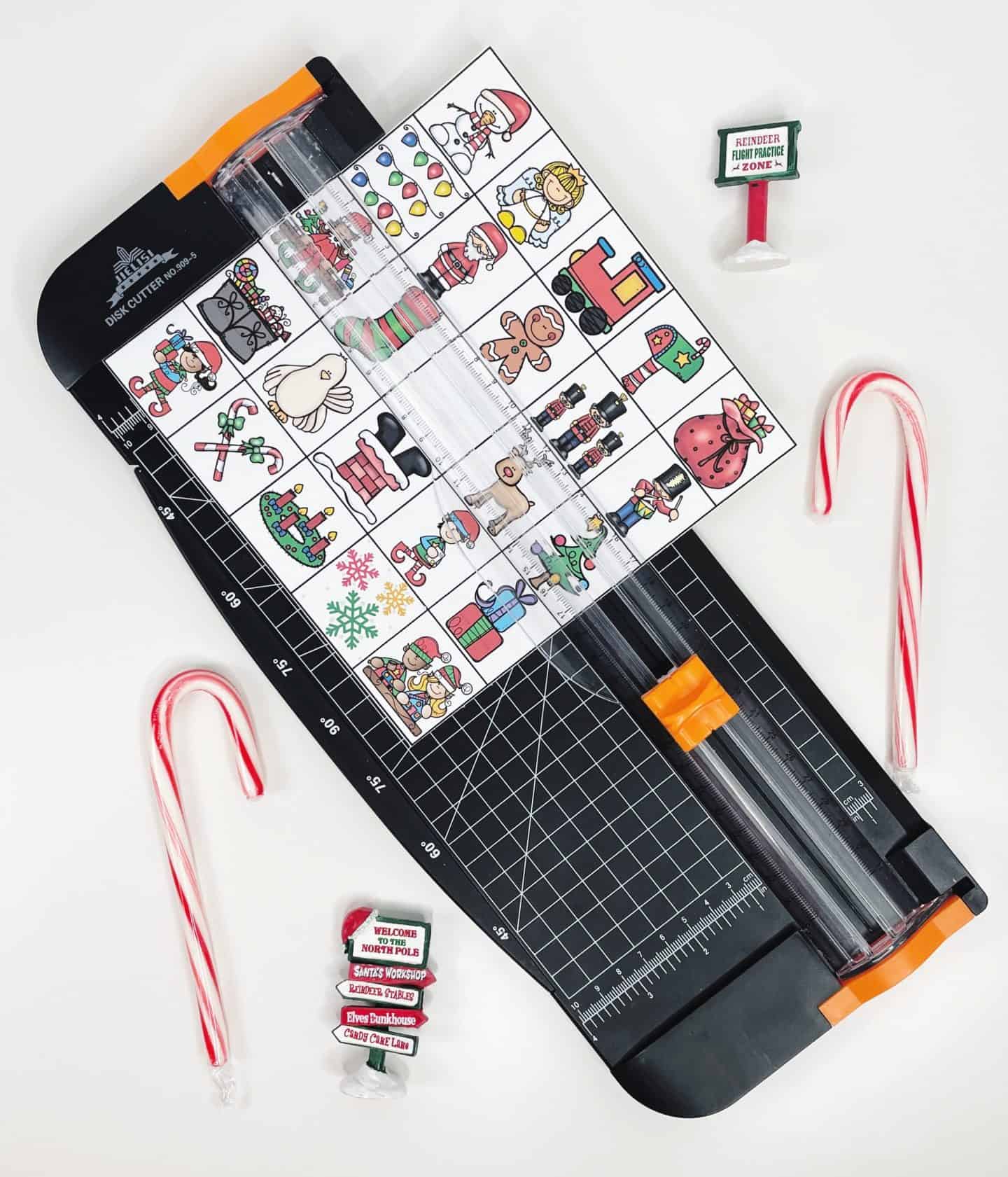 A Christmas Bingo calling card sheet is placed on a paper cutter, partially sliced into smaller squares. The card features colorful holiday illustrations like Santa, ornaments, gifts, and candy canes. Surrounding the setup are festive items including two candy canes and mini North Pole signs that read phrases like “Welcome to the North Pole” and “Reindeer Flight Practice Zone.” The scene captures a holiday craft in progress.