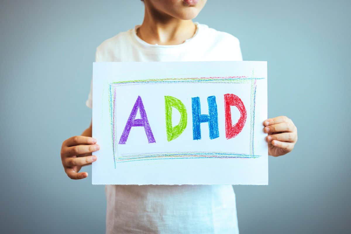 A child holding a white sign with "ADHD" written in large, colorful letters against a plain background. The letters "A," "D," "H," and "D" are in red, green, blue, and purple, respectively.